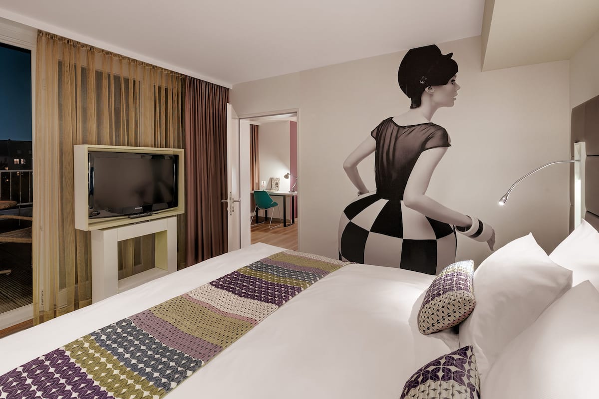 A hotel room with a large bed, a television on a stand, a balcony with a table and chairs, and a view of a cityscape at night.