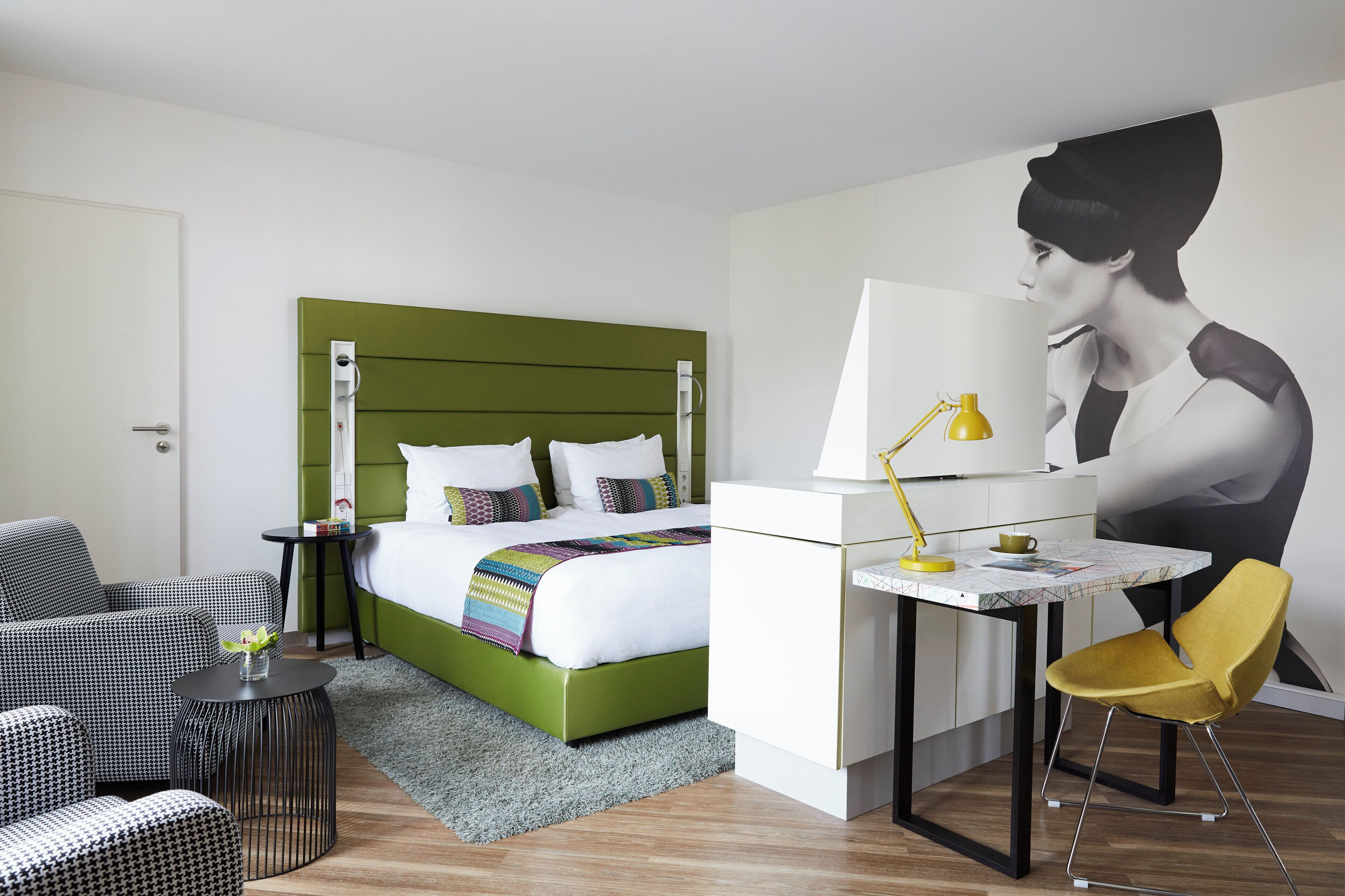 Habitación de hotel con una cama verde, un escritorio con lámpara, una silla y un mural grande en la pared.