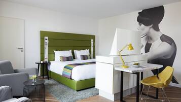 Habitación de hotel con una cama verde, un escritorio con lámpara, una silla y un mural grande en la pared.