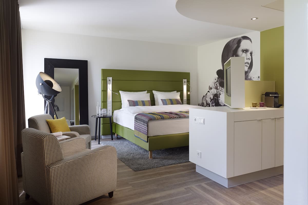 A modern hotel room with a green headboard bed, a small side table with a lamp, a sofa, and a large mirror.