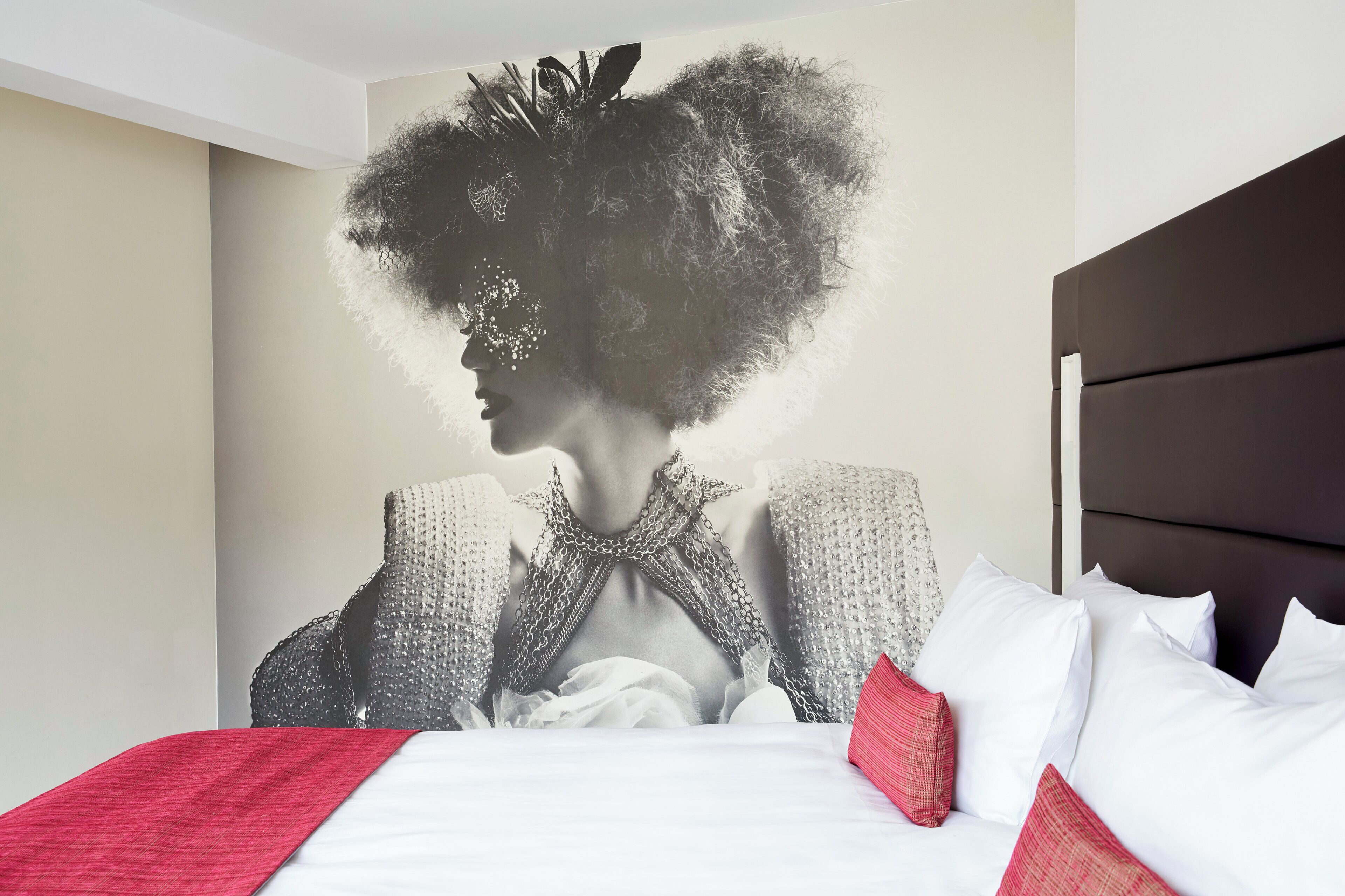 Una cama con ropa de cama blanca y roja, un mural blanco y negro en la pared y un cabecero oscuro.