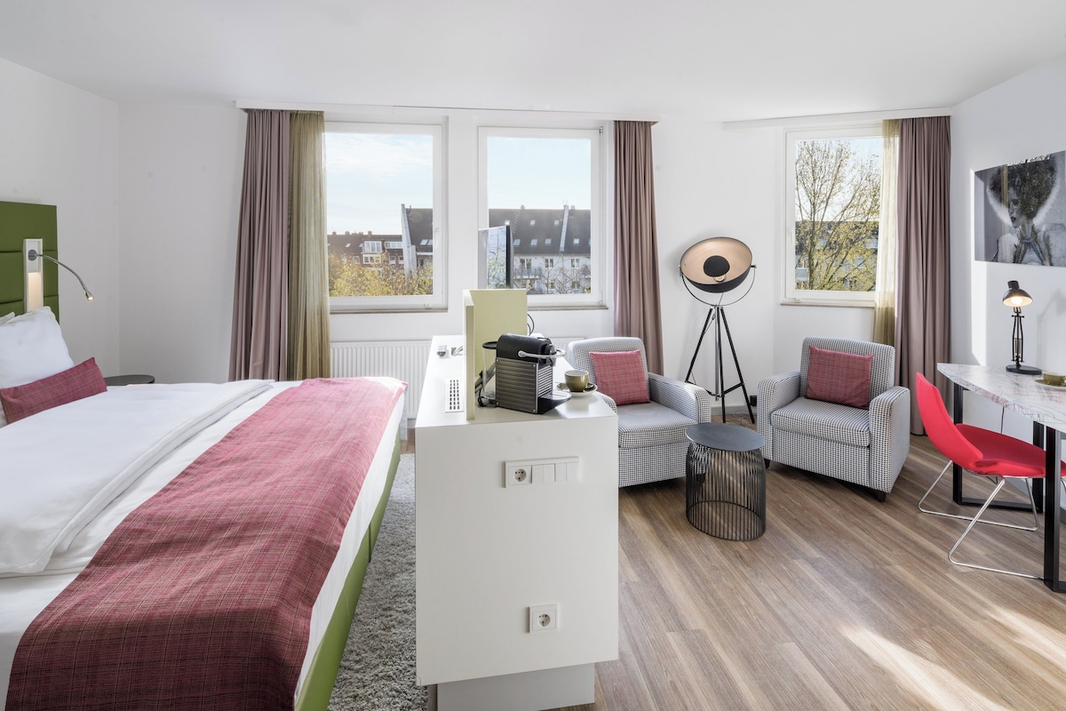 Hotelkamer met een bed, bureau, twee stoelen, een tafeltje en uitzicht op gebouwen door de ramen.