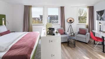 A hotel room with a bed, desk, two chairs, a small table, and a view of buildings through the windows.