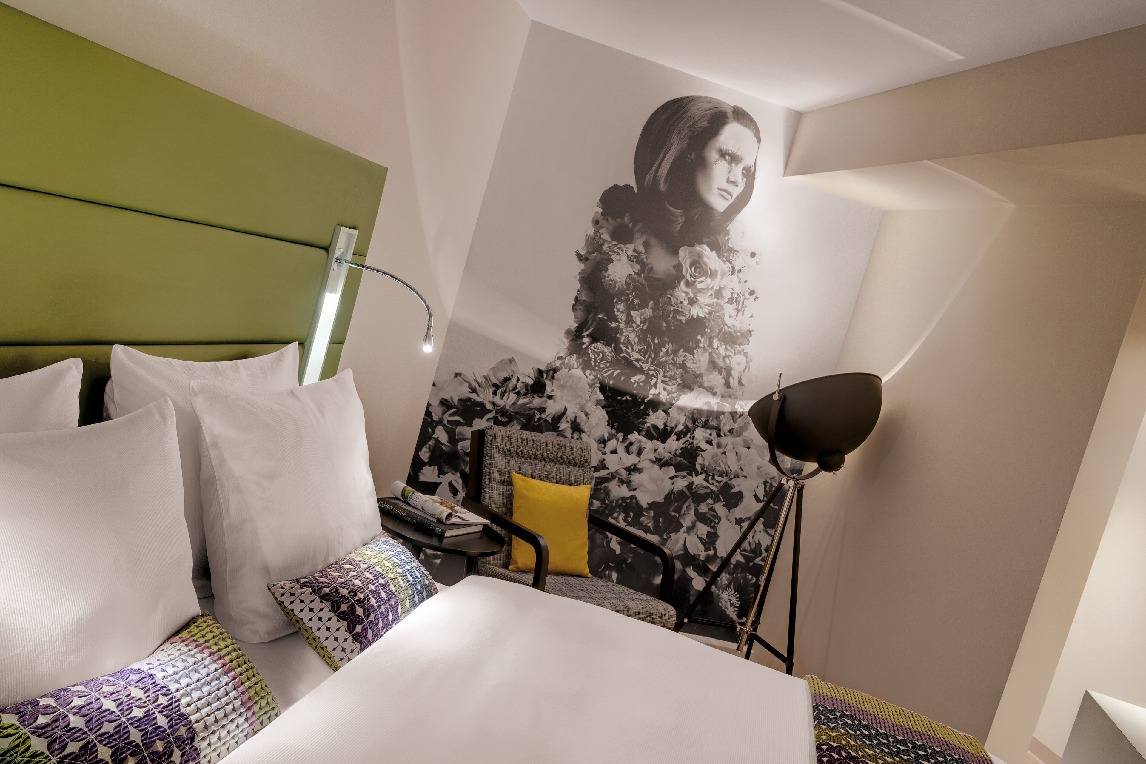A hotel room with a bed, a chair with a yellow pillow, a desk with books, and a large floral wall mural.