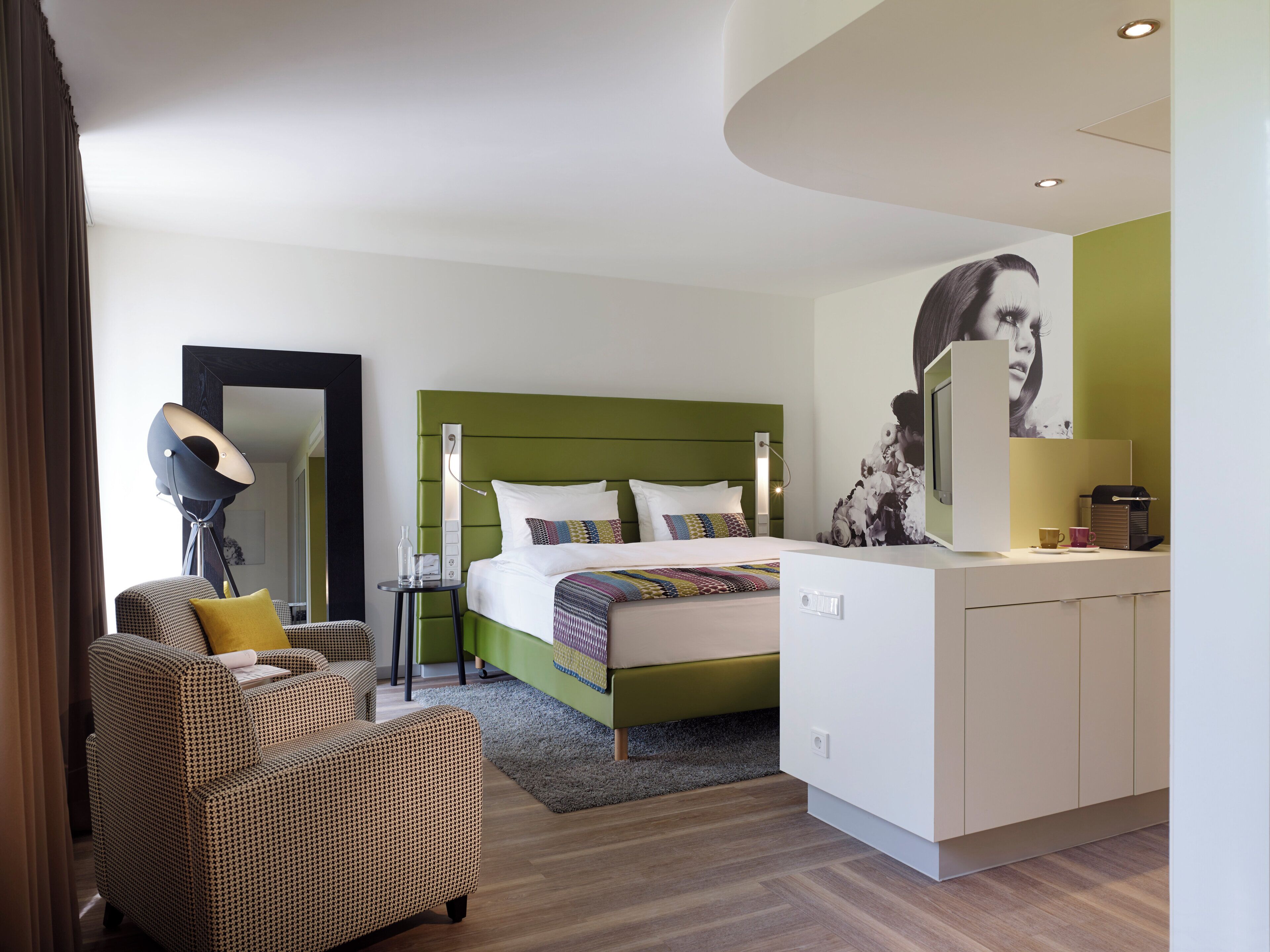 A modern hotel room with a green headboard, a bed with multiple pillows, a bedside table with a lamp, a chair, and a large mirror.