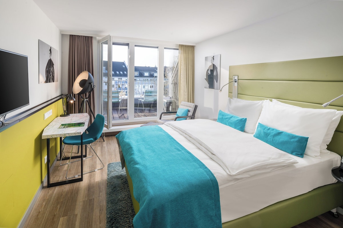 Een hotelkamer met een bed, een bureau, een stoel, een televisie en een balkon met uitzicht op gebouwen.