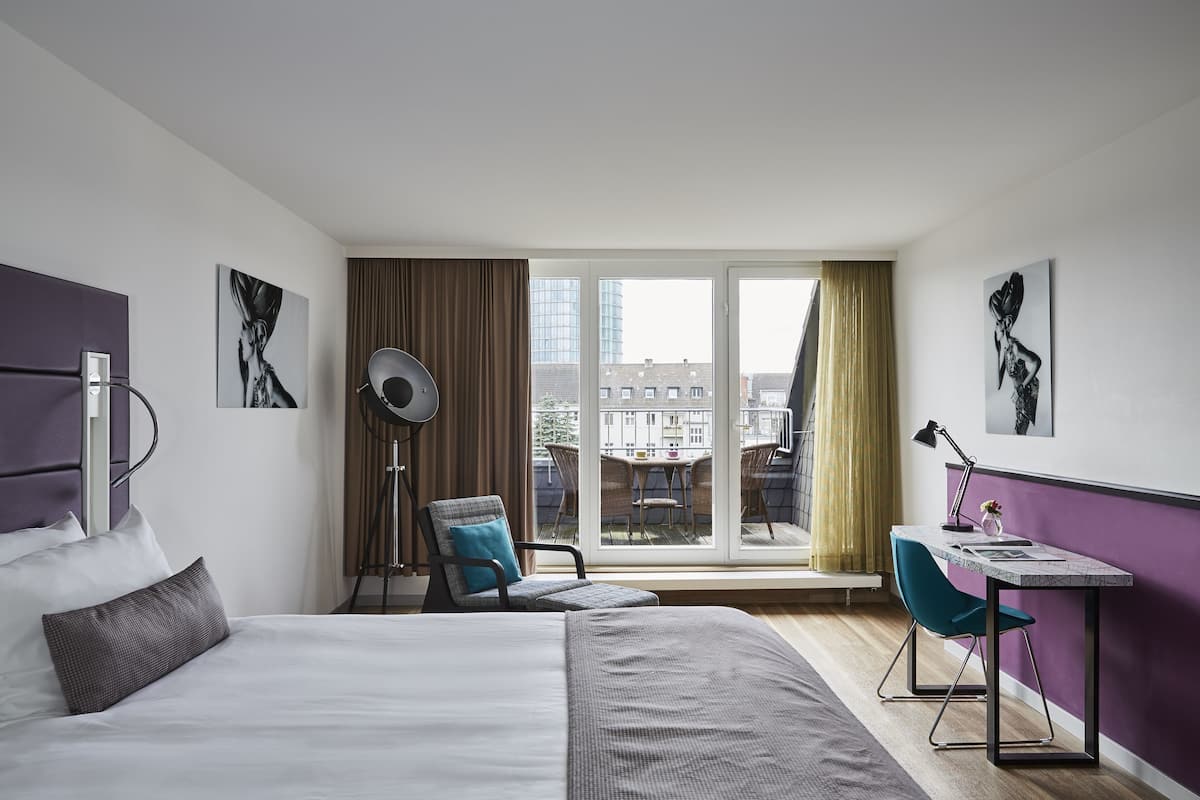 A modern hotel room with a large bed, a desk with a chair, a window with a view of buildings, and artwork on the walls.