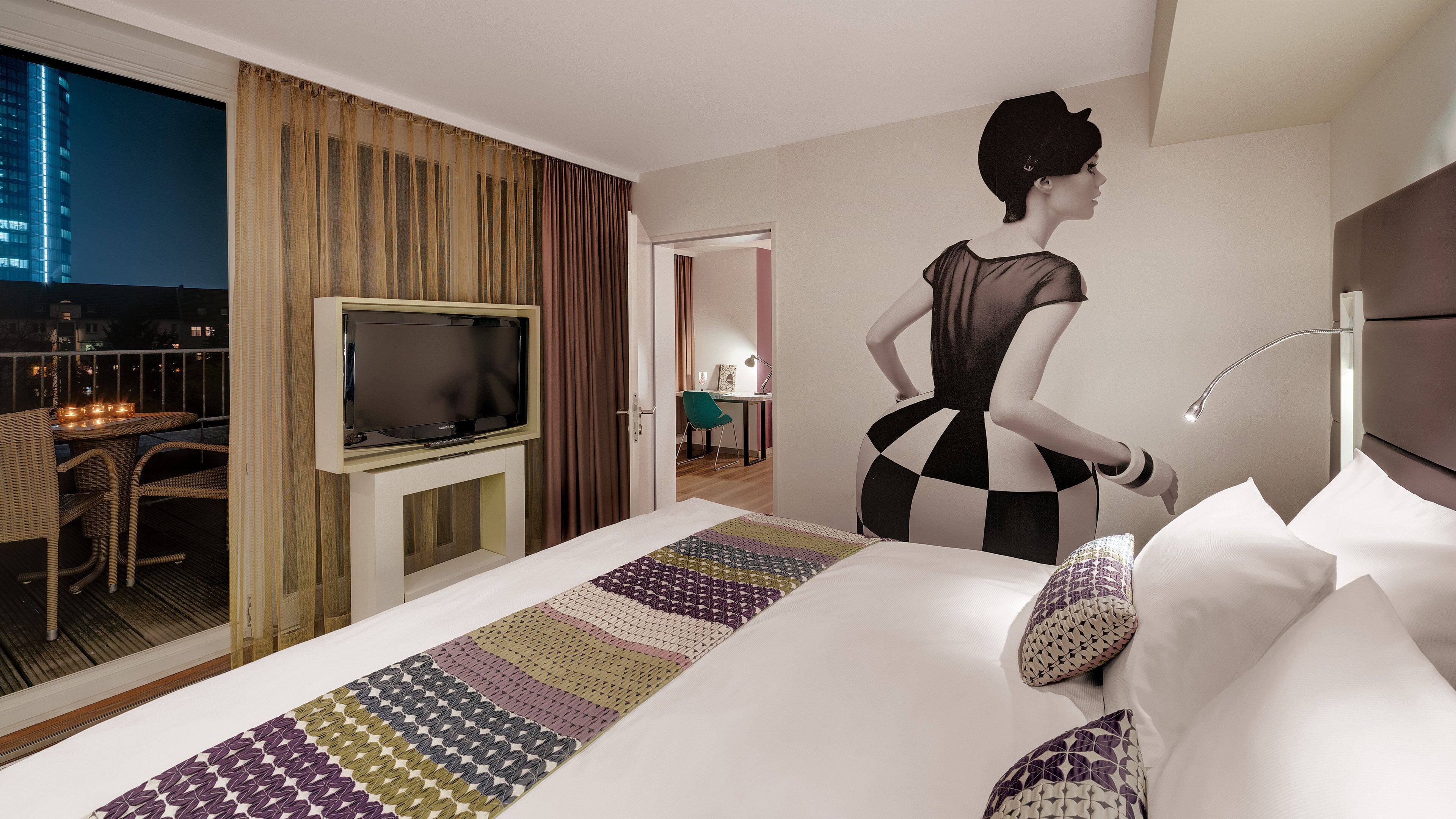 A hotel room with a bed, a television, a desk, and a view of the city at night.