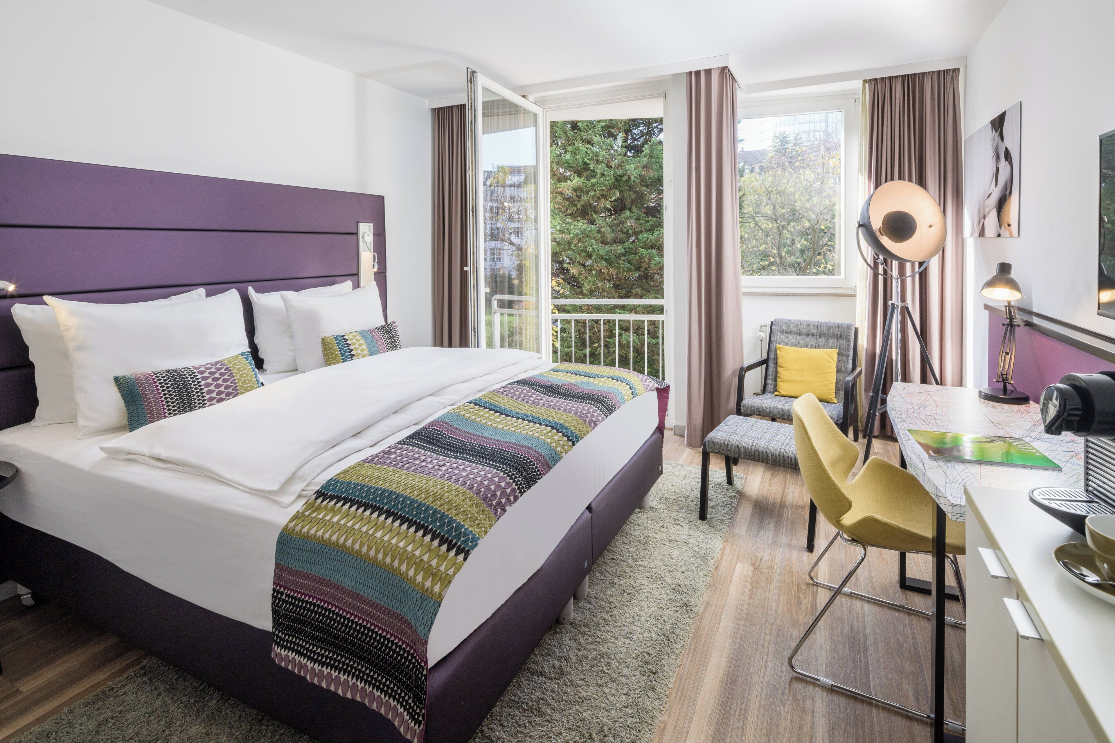 Una habitación de hotel con cama, escritorio, silla y vistas a árboles por la ventana.