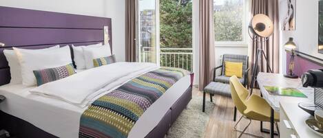 A hotel room with a bed, a desk, a chair, and a view of trees through the window.
