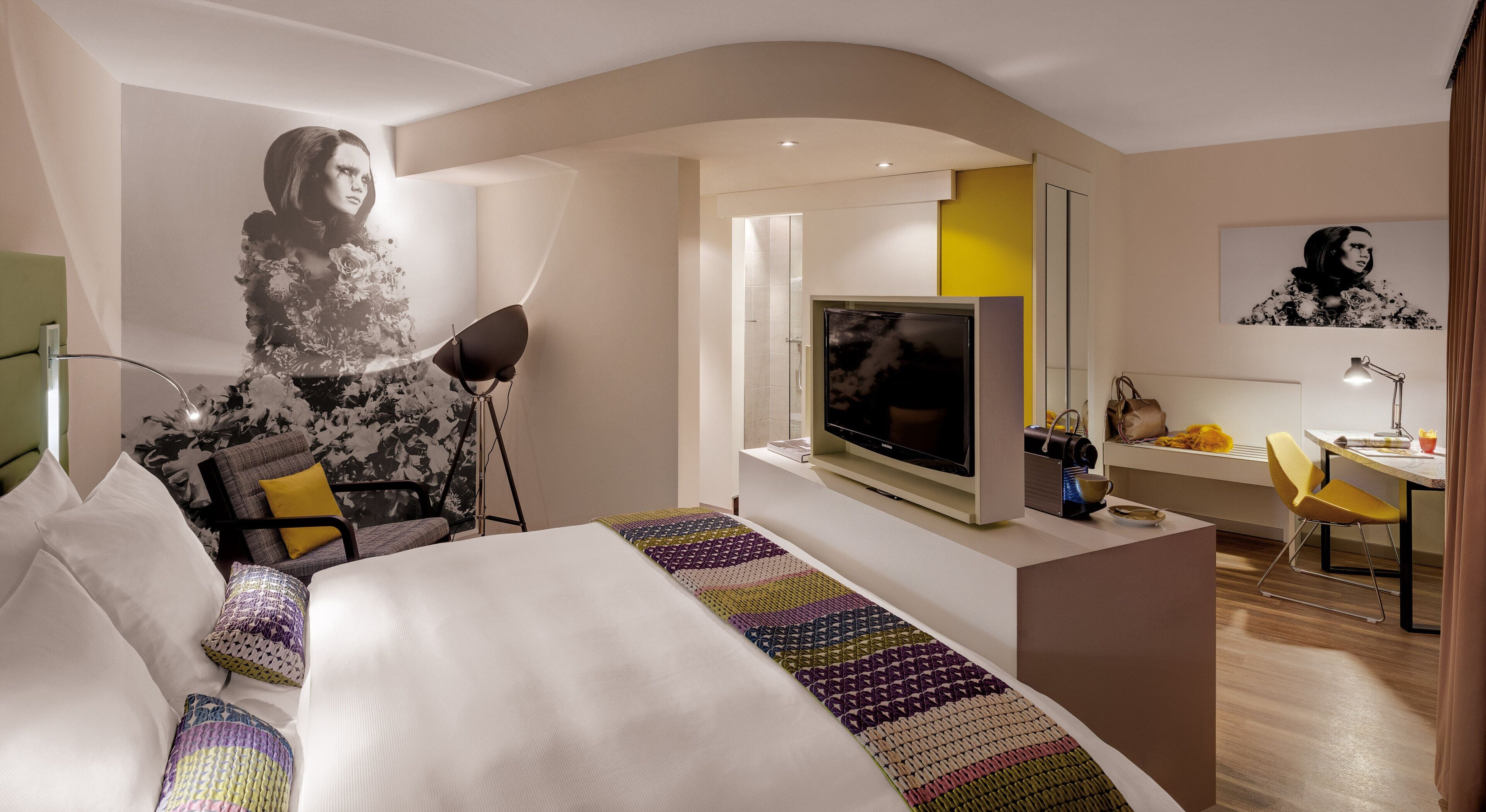 A modern hotel room with a large bed, a flat-screen TV, a desk with a chair, and a large wall mural.