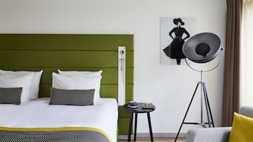 A hotel room with a green headboard, a bed with white and black bedding, a small round table with a cup and books, and a floor lamp.