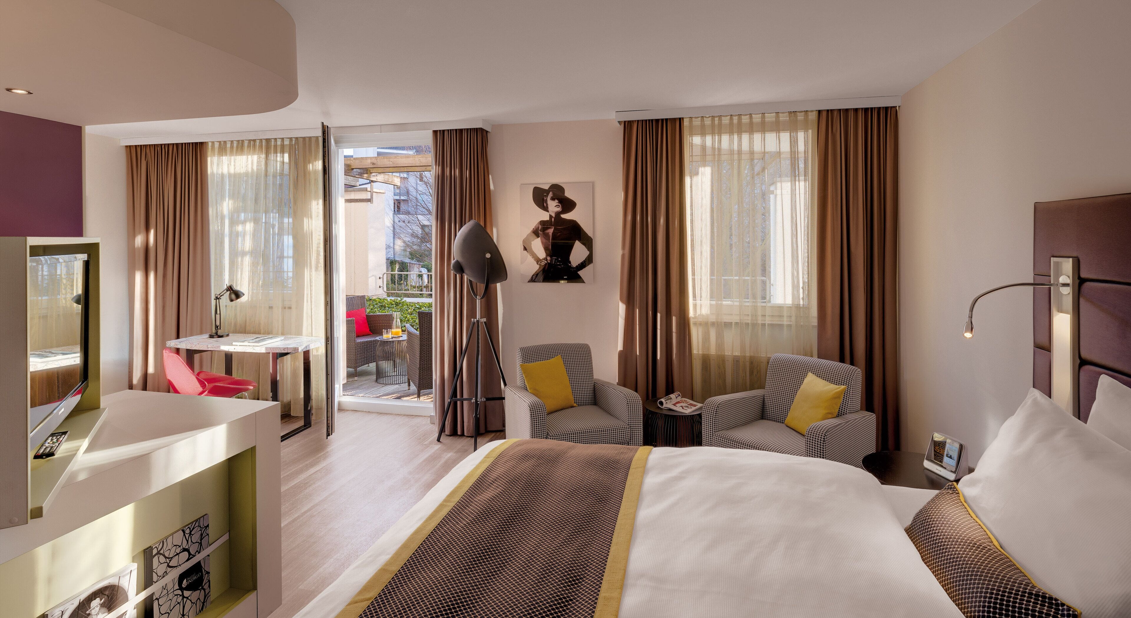 A hotel room with a bed, two chairs, a desk, a TV, and a balcony with outdoor seating.