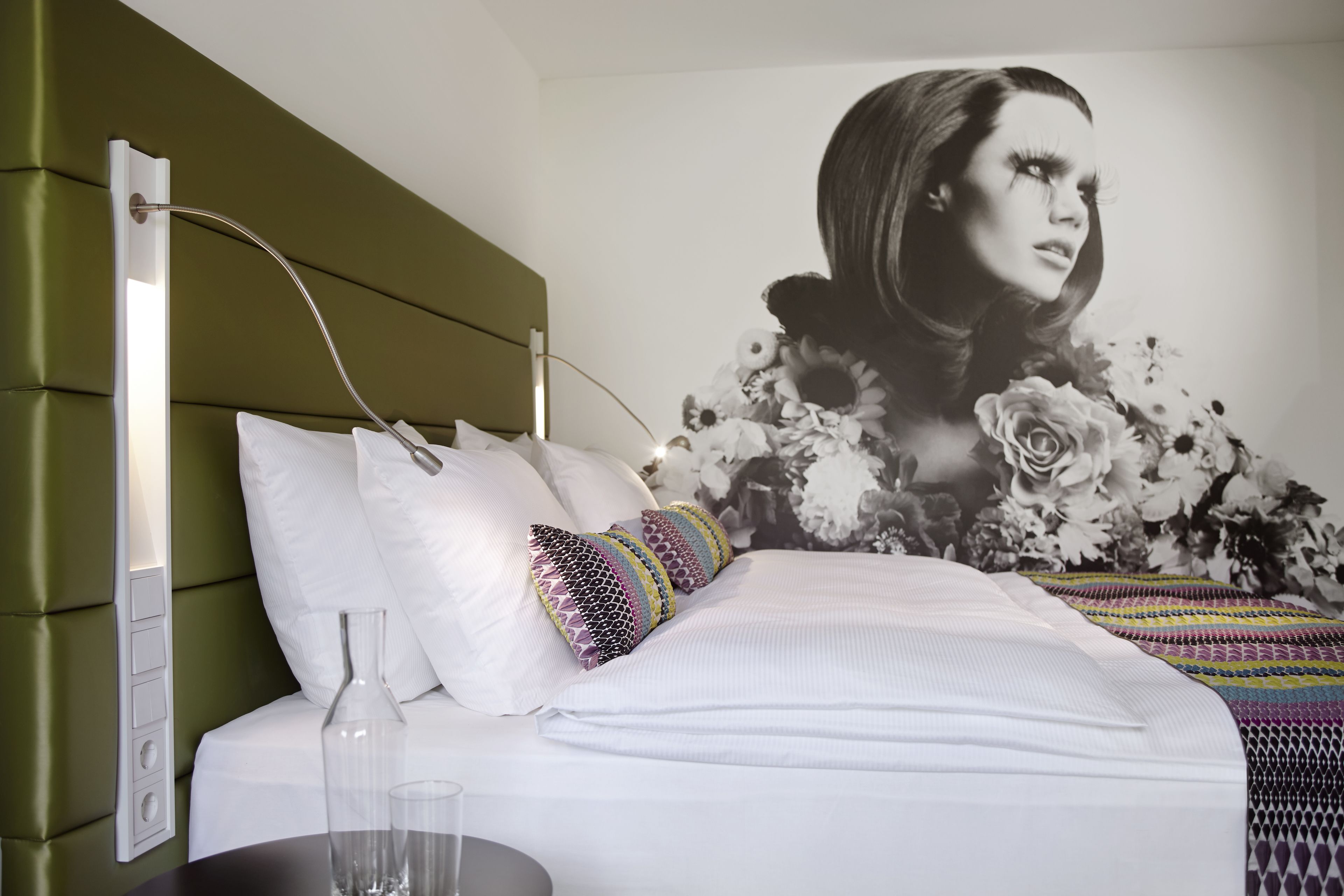 Una habitación de hotel moderna con cabecera verde, ropa de cama blanca y un gran mural floral en la pared.