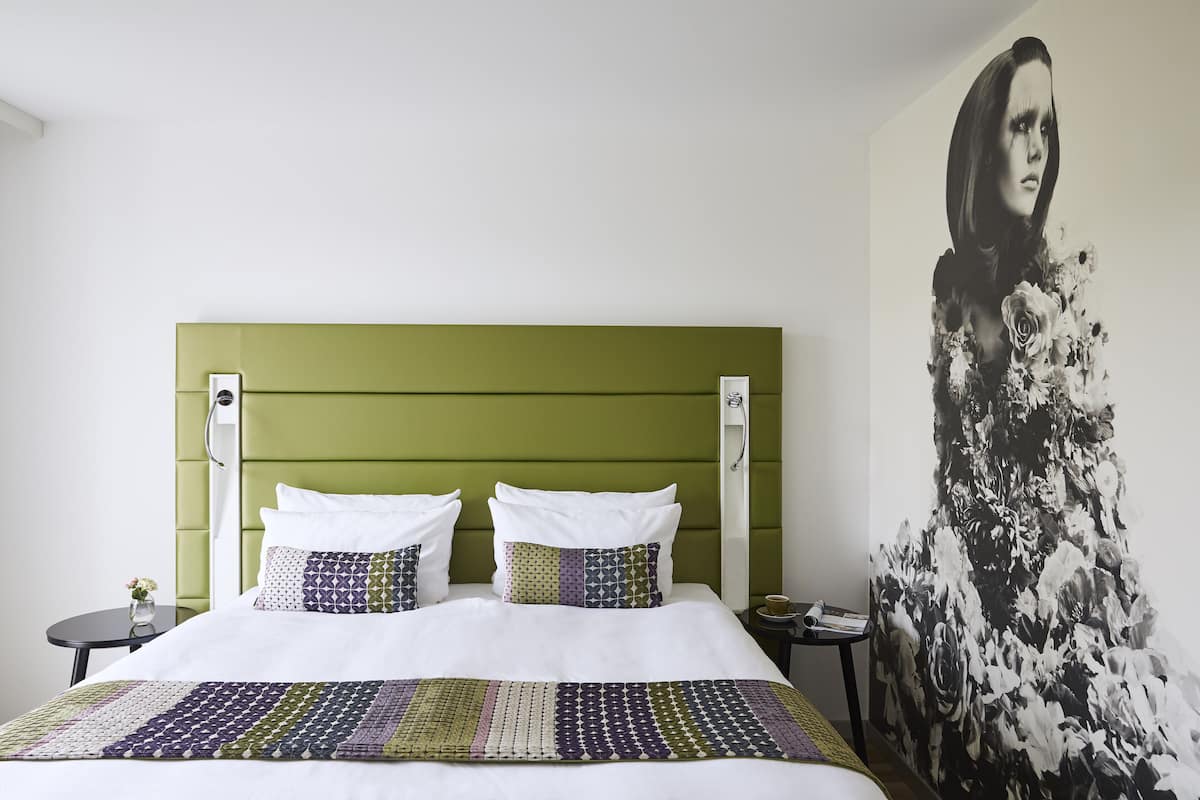 A bed with a green headboard, white bedding, and patterned pillows. A floral artwork is on the wall to the right.