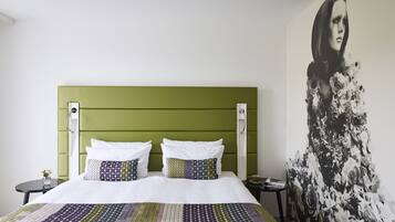 A bed with a green headboard, white bedding, and patterned pillows. A floral artwork is on the wall to the right.