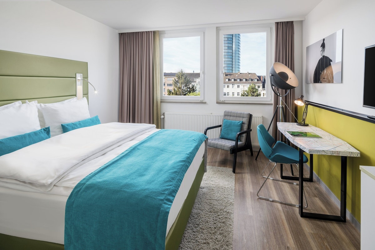 Een hotelkamer met een bed, een bureau, een stoel en uitzicht op gebouwen door het raam.