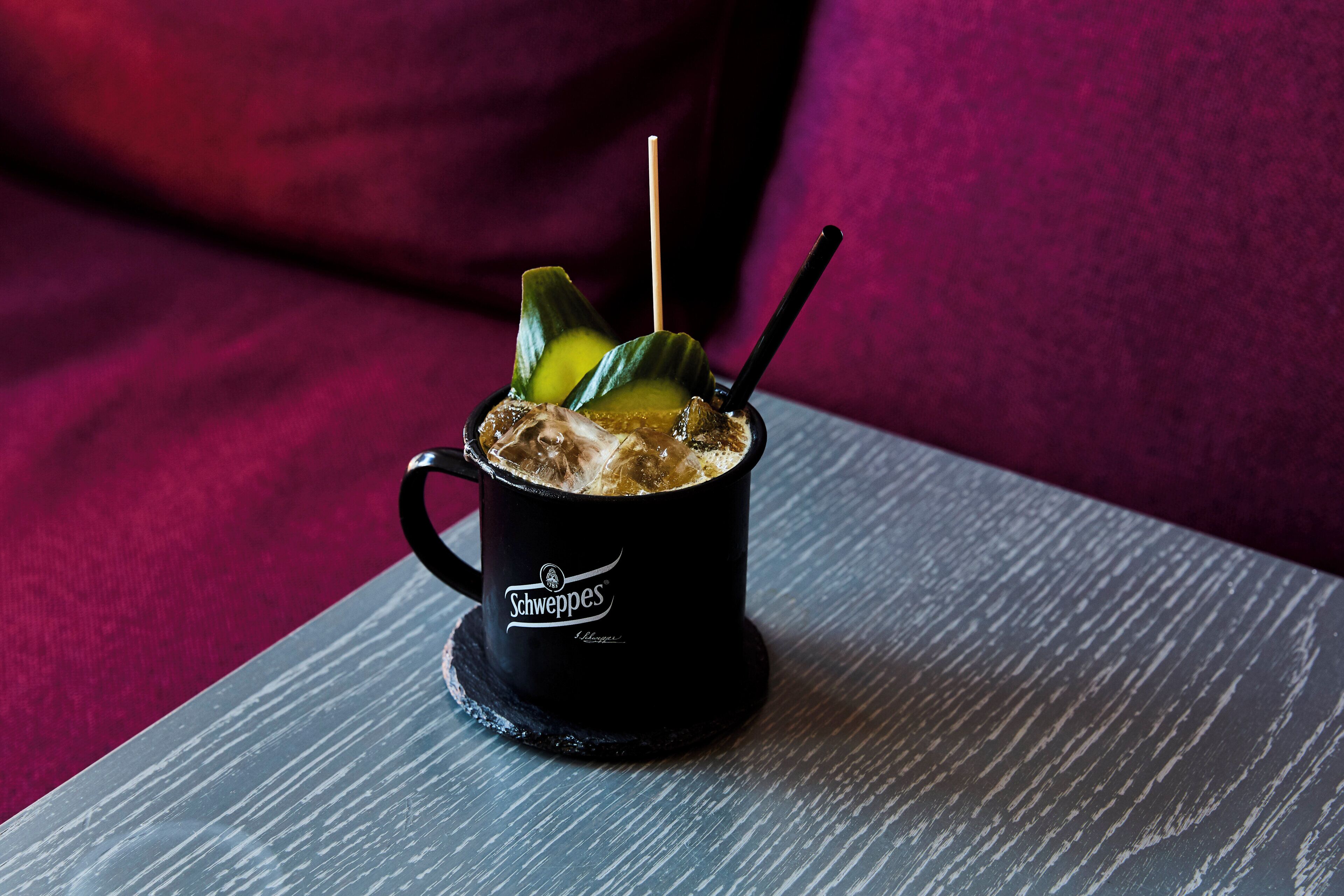 Une tasse noire avec une paille et une tranche de citron vert, posée sur un dessous de verre, elle-même placée sur une table grise.