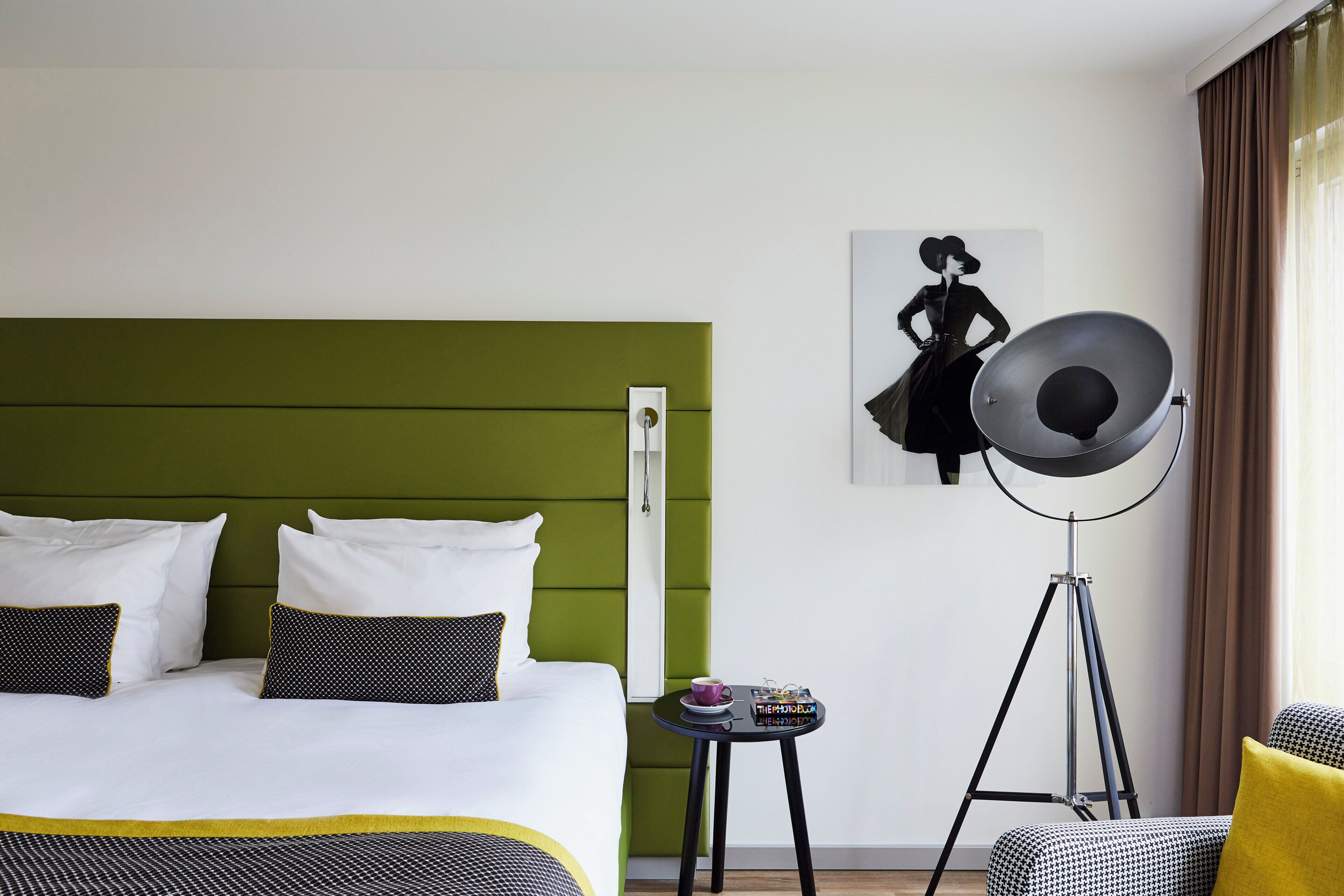 Quarto de hotel com uma cama, uma mesinha com uma xícara e uma revista, um abajur e um quadro na parede.