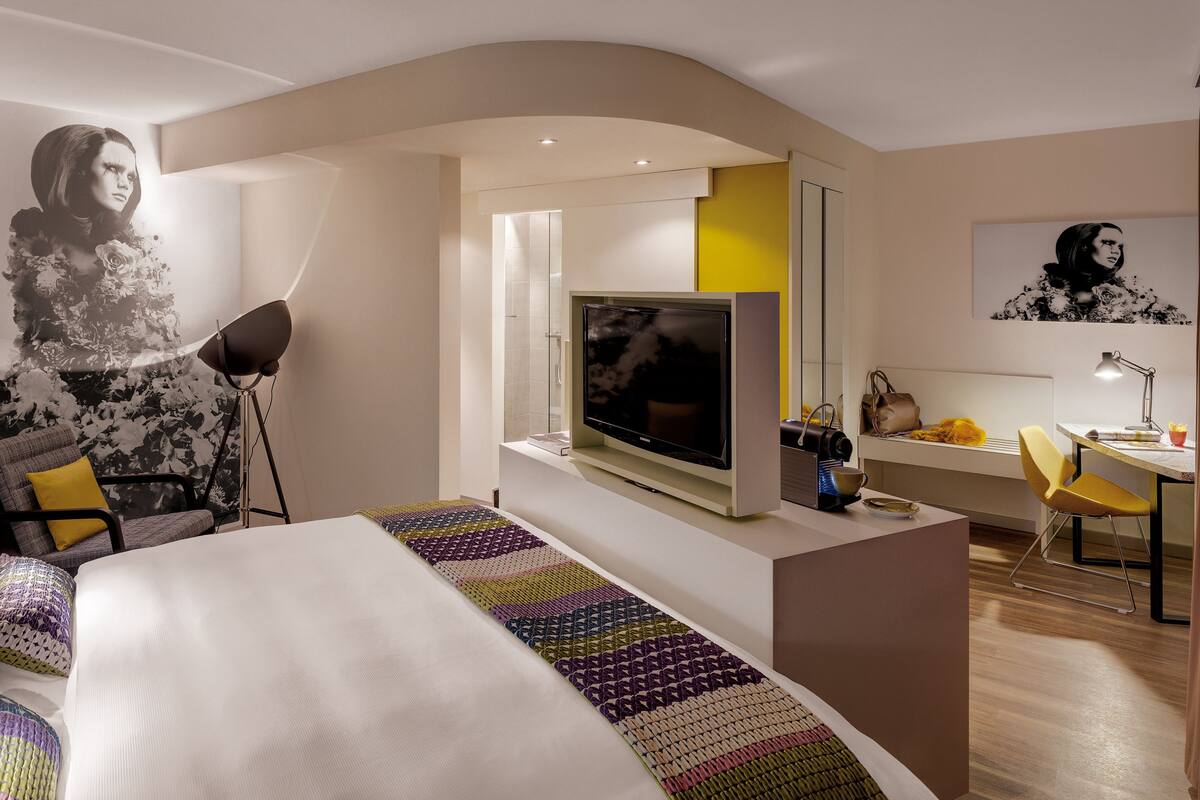 A modern hotel room with a large bed, a flat-screen TV, a desk with a chair, and a large window.