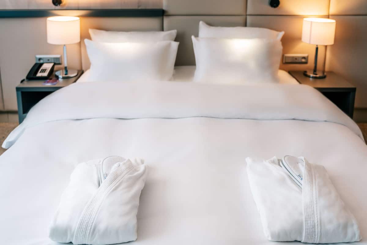 A hotel room with a neatly made bed, two bedside lamps, a nightstand with a phone, and two towels on the bed.