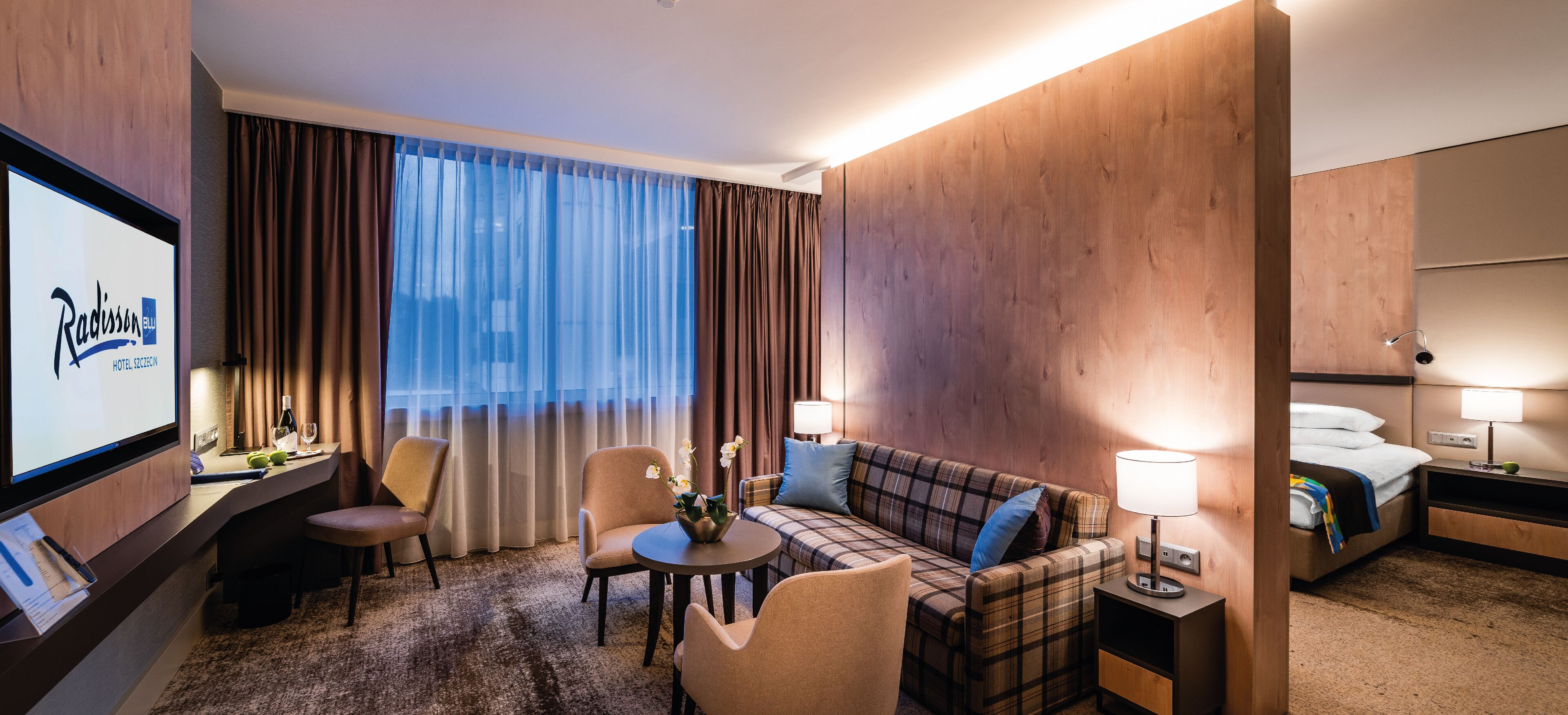 A hotel room with a large flat-screen TV, a sofa, a small round table, two armchairs, a bed, and a nightstand with a lamp.