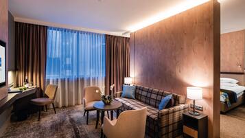 A hotel room with a large flat-screen TV, a sofa, a small round table, two armchairs, a bed, and a nightstand with a lamp.