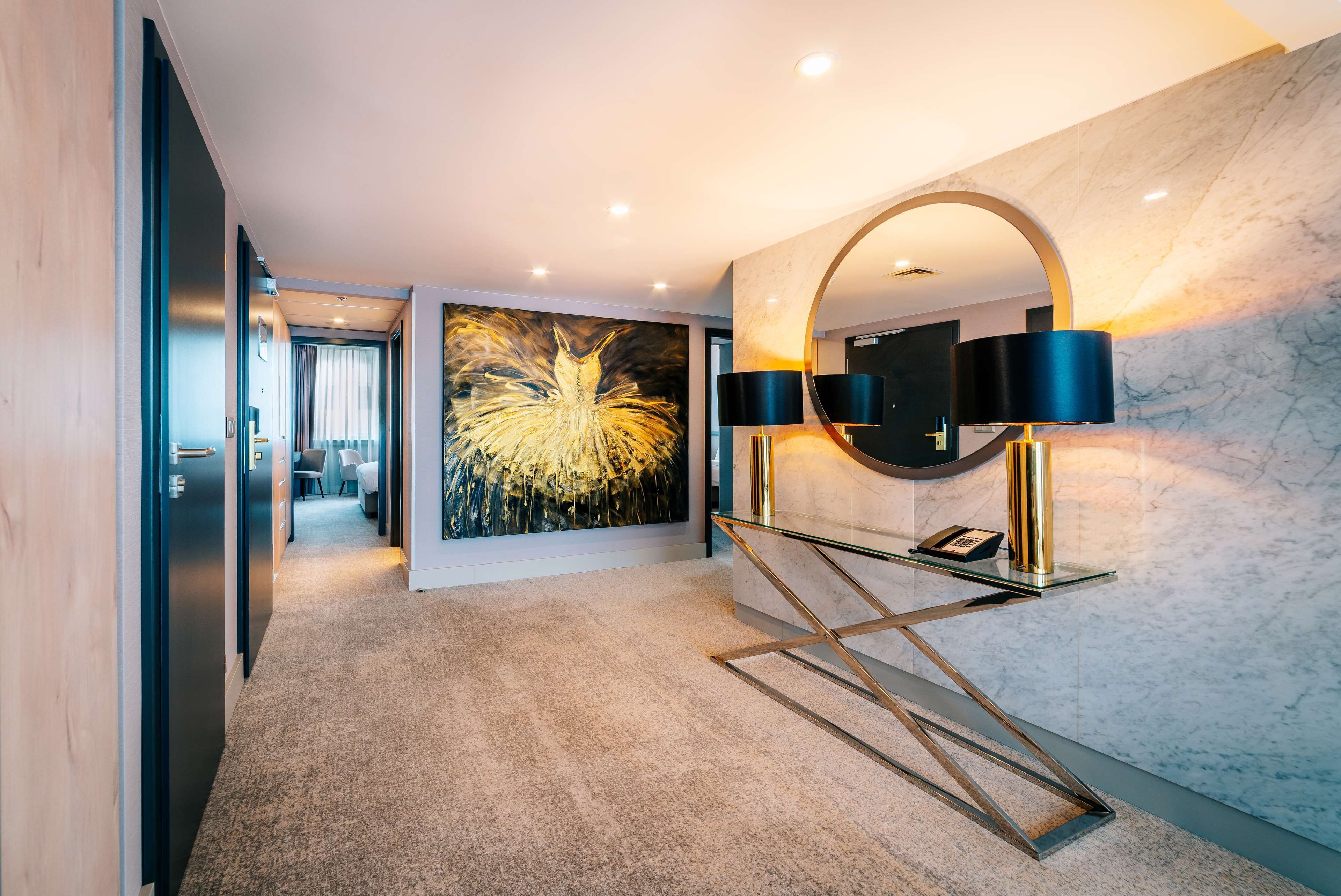 Un hall d’hôtel moderne avec un mur de marbre, une grande peinture abstraite, une console en verre avec un téléphone et un miroir rond.