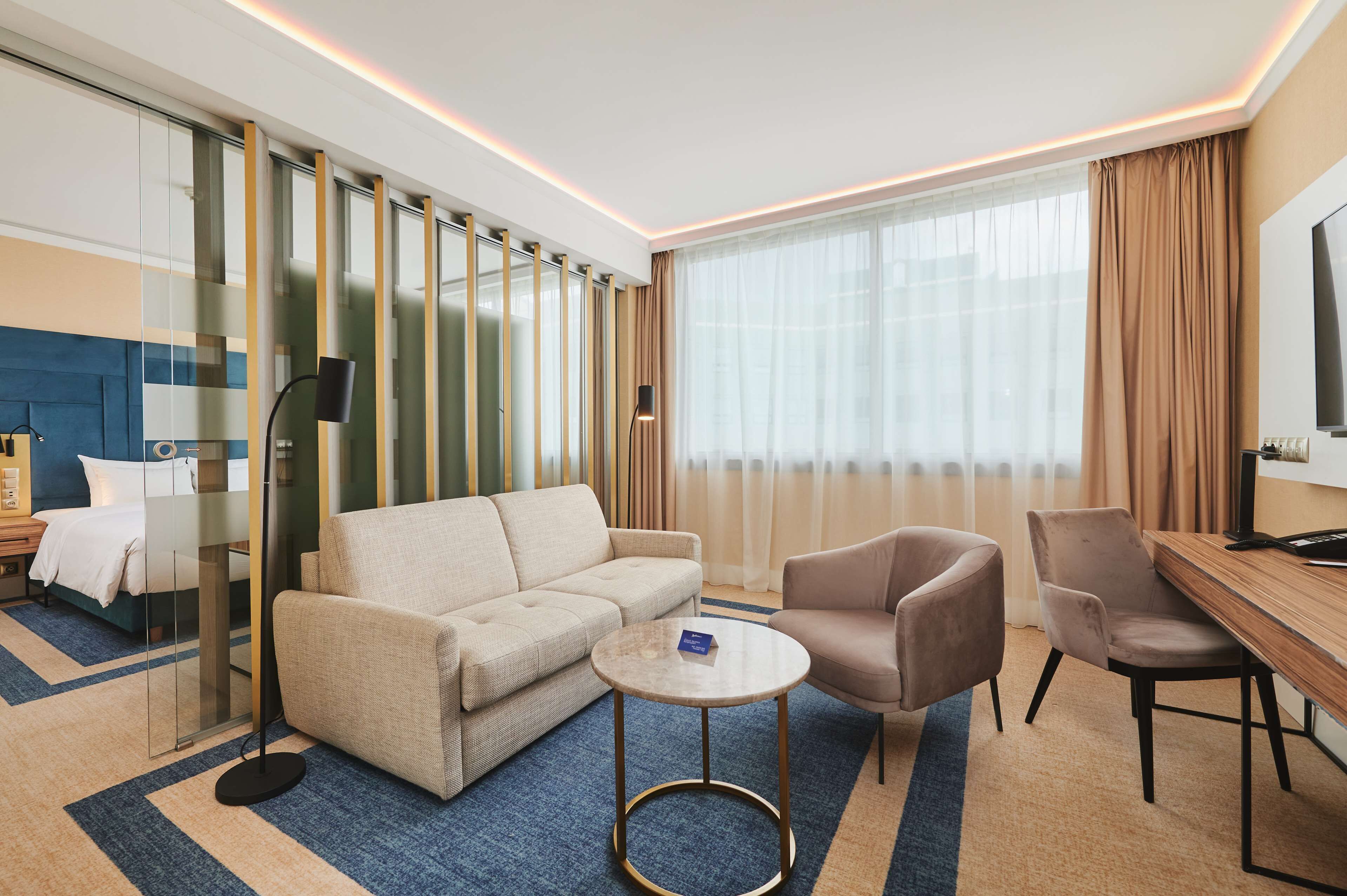 Una habitación de hotel con sofá, sillón, una mesita redonda y televisión sobre un mueble.