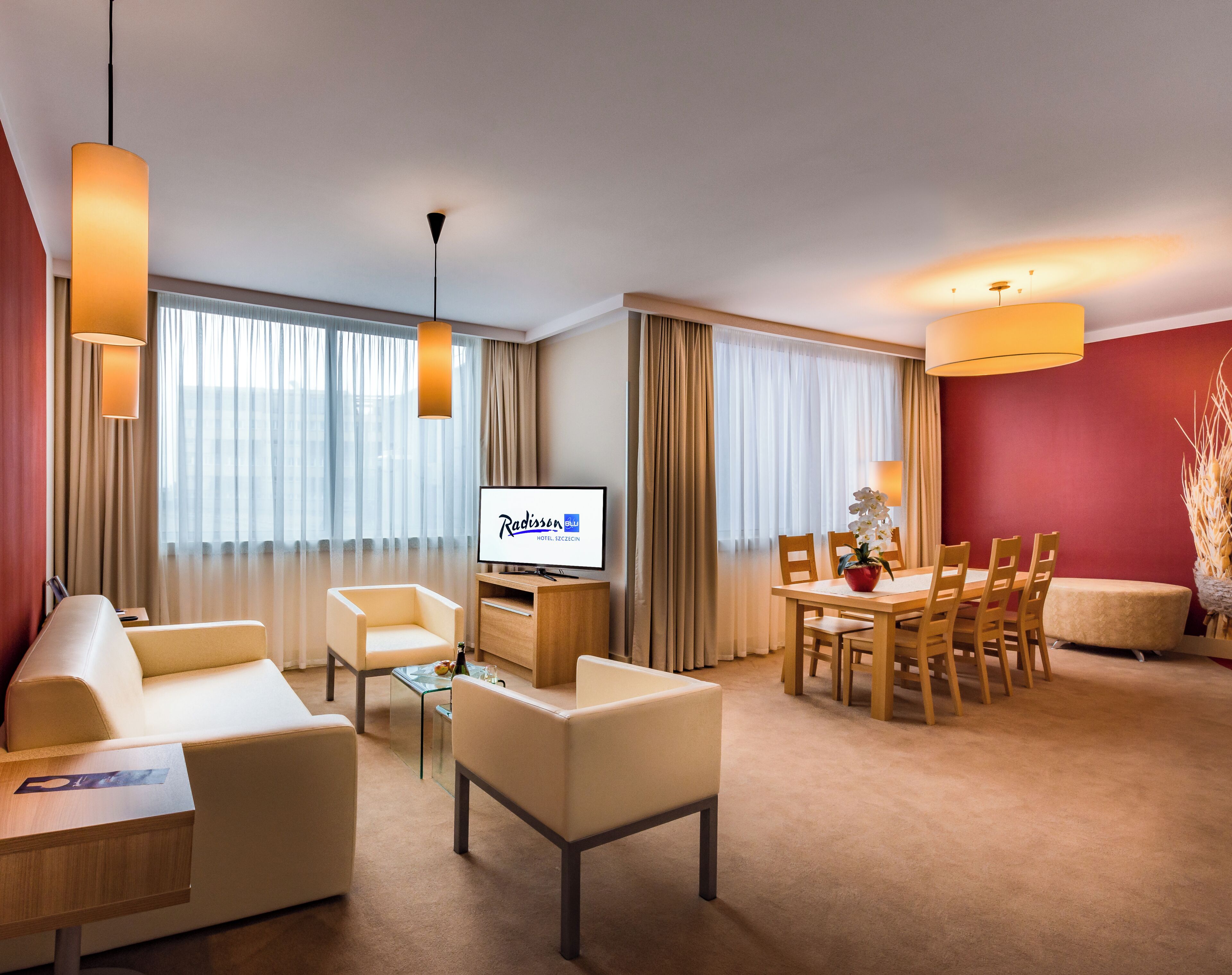 Habitación de hotel con sofá, sillas, una mesa de comedor, televisor y una pared de acento roja.