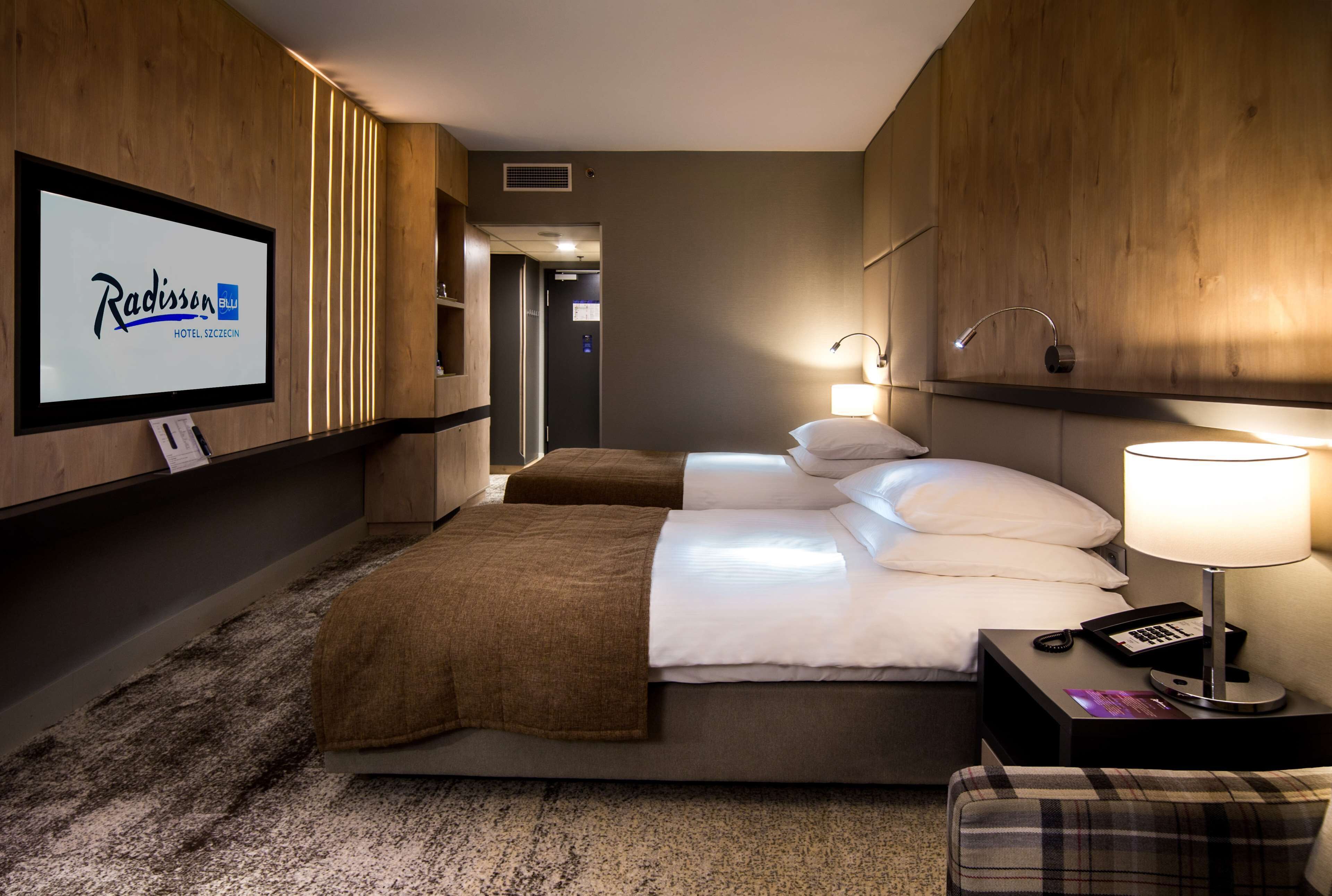 A hotel room with a large flat-screen TV displaying Radisson Blu Hotel logo, a bed with white linens, a brown throw blanket, a nightstand with a lamp, and a plaid chair.