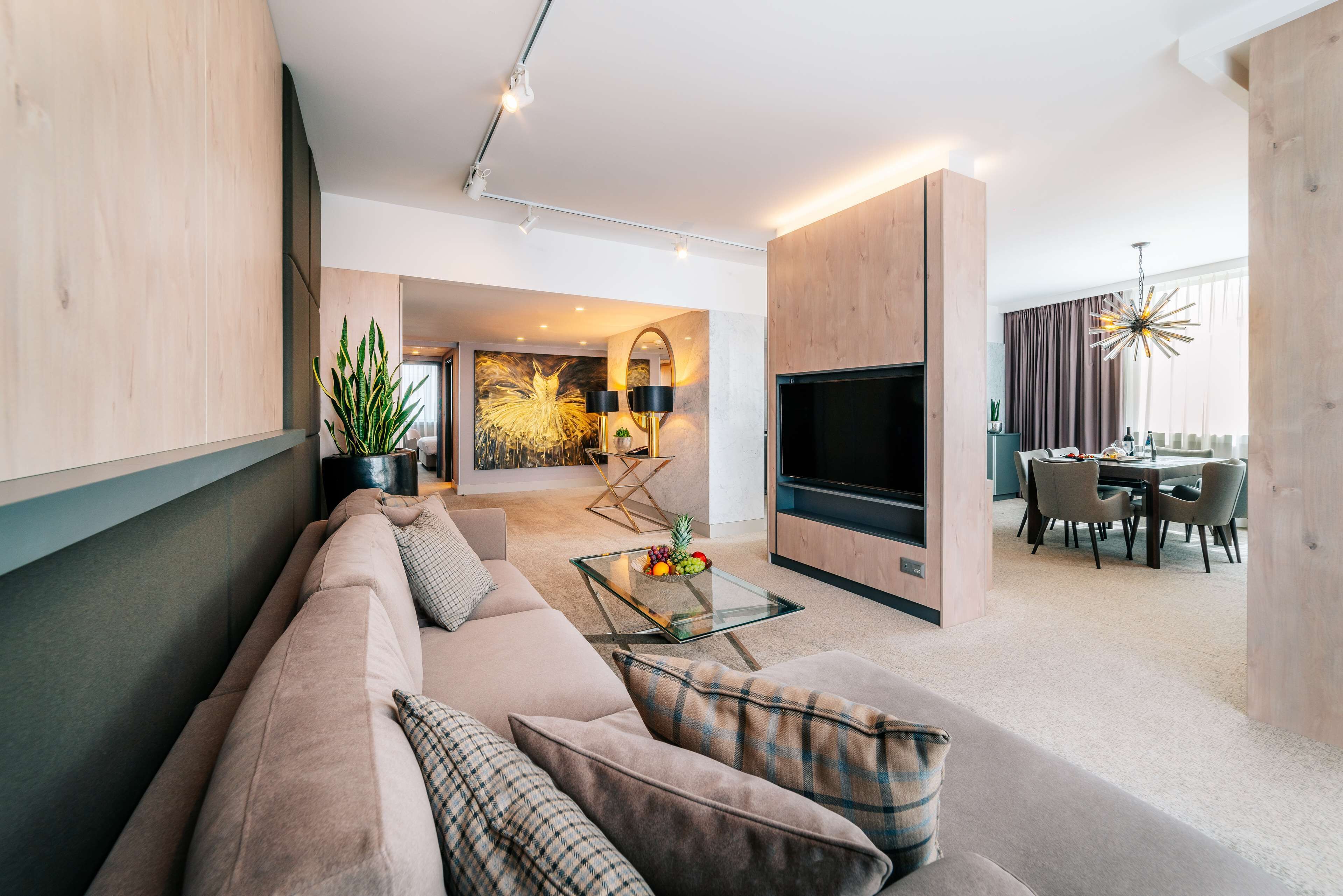 Ein modernes Wohnzimmer mit einem beigefarbenen Sofa, einem Glastisch mit Obst, einem Flachbildfernseher und einem Essbereich mit einer Kronleuchter.