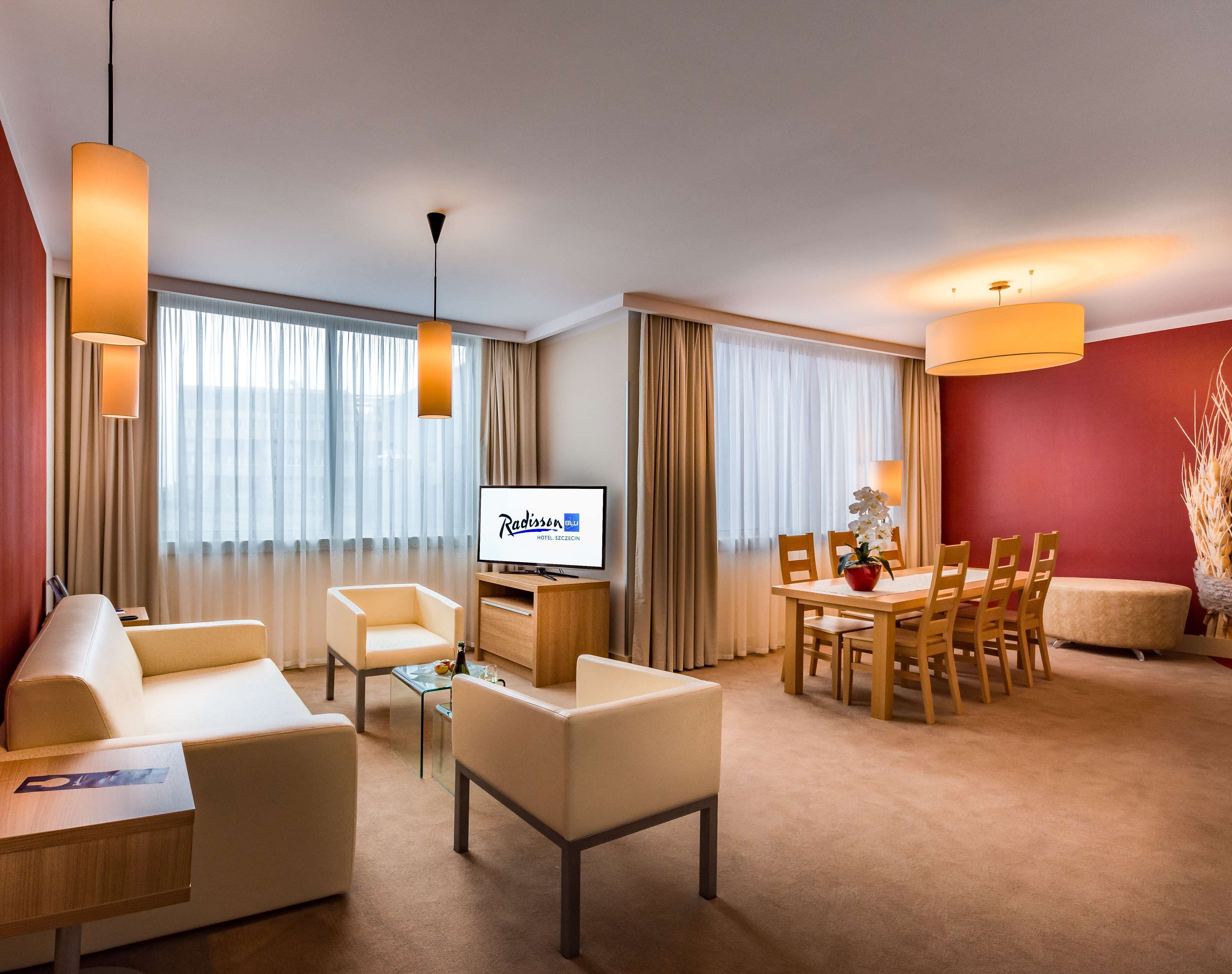 A hotel room with a sofa, chairs, a dining table, a TV, and a large window with curtains.