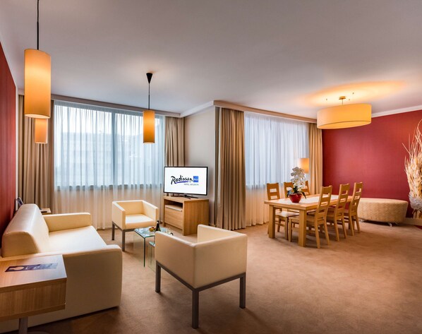 A hotel room with a sofa, chairs, a dining table, a TV, and a large window with curtains.