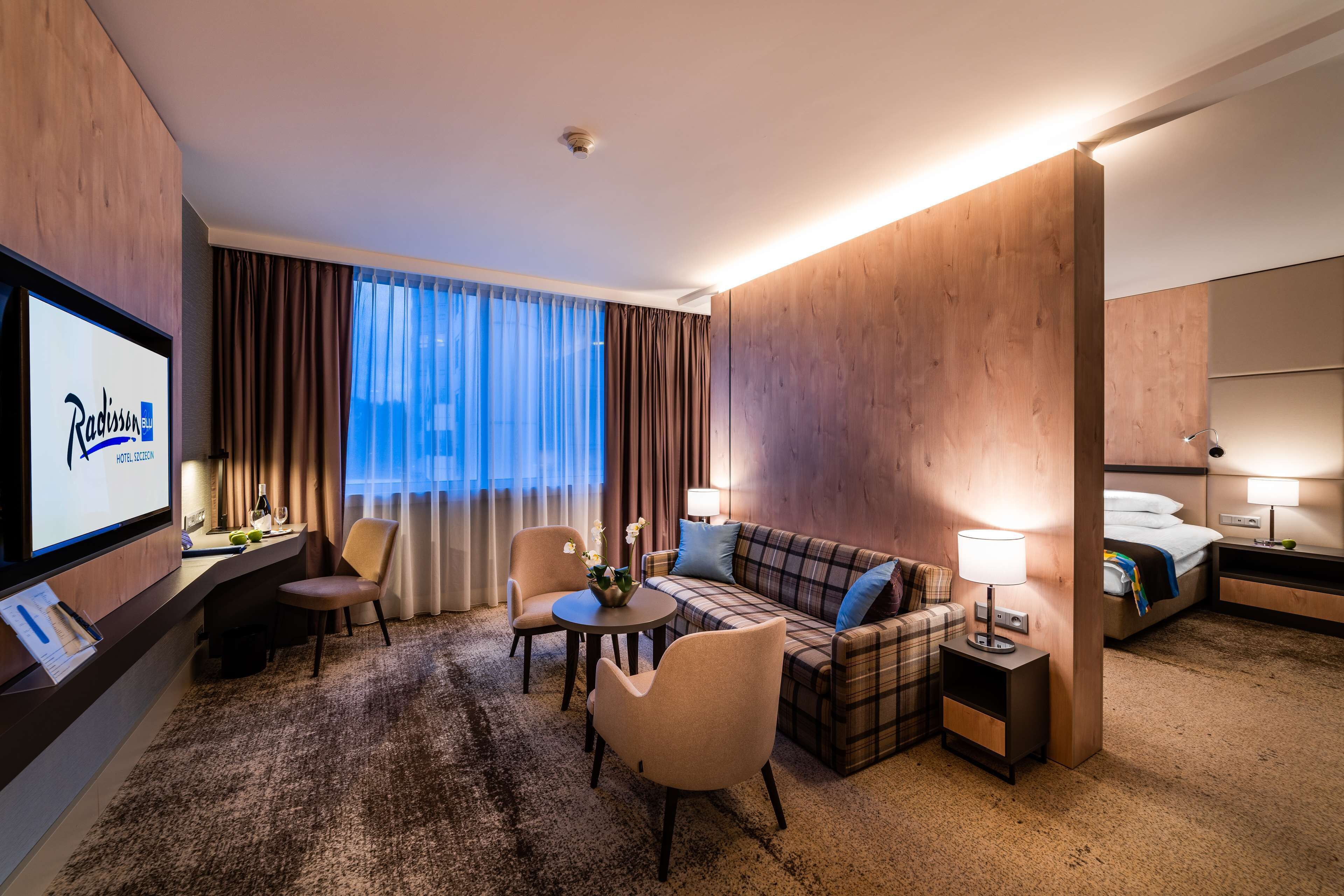 A hotel room with a TV, a sofa, a chair, a small table, a lamp, and a bed.