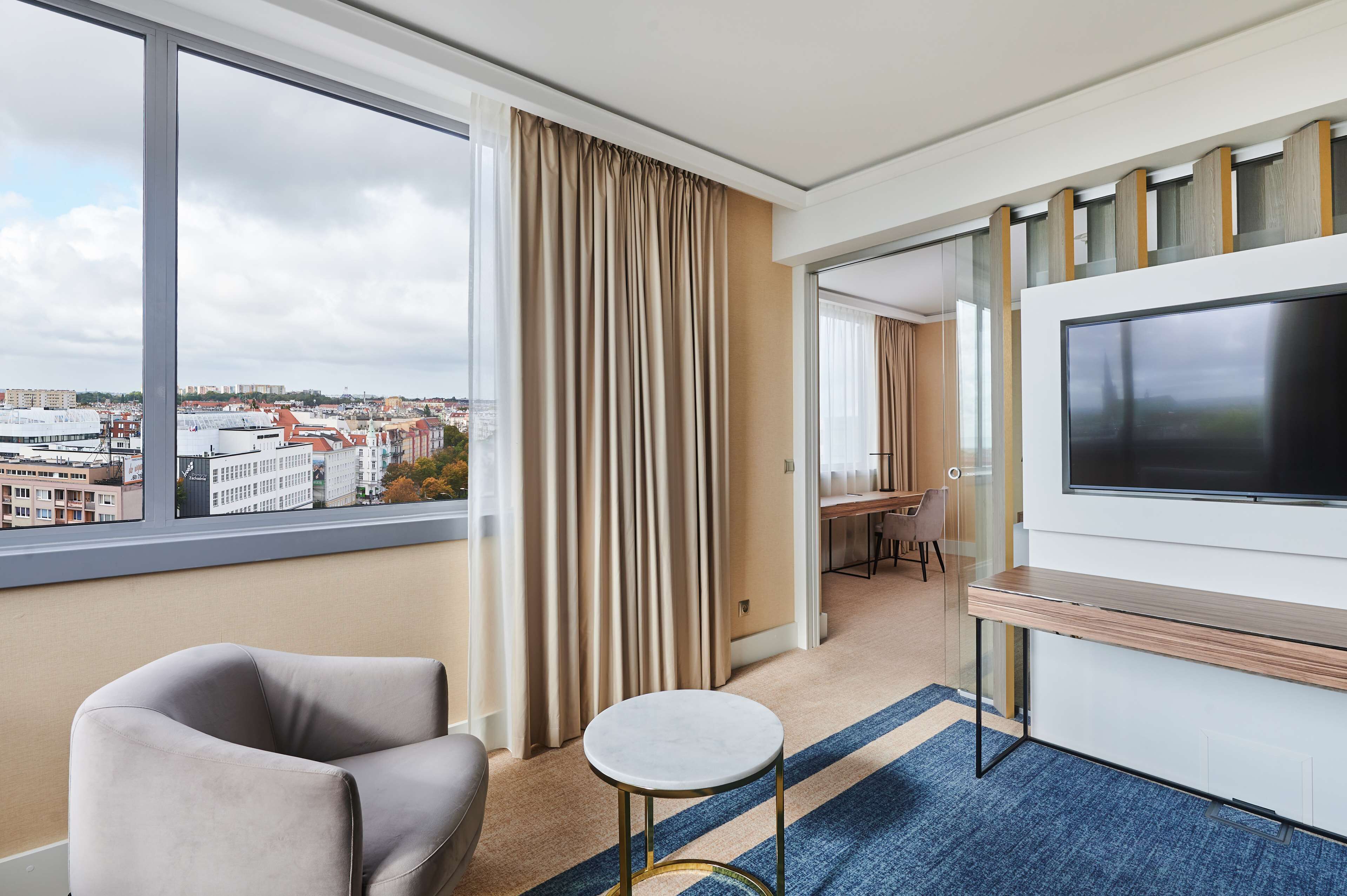 A hotel room with a large window, a flat-screen TV, a sofa, a small round table, and a desk with chairs.