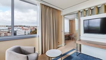 A hotel room with a large window, a flat-screen TV, a sofa, a small round table, and a desk with chairs.