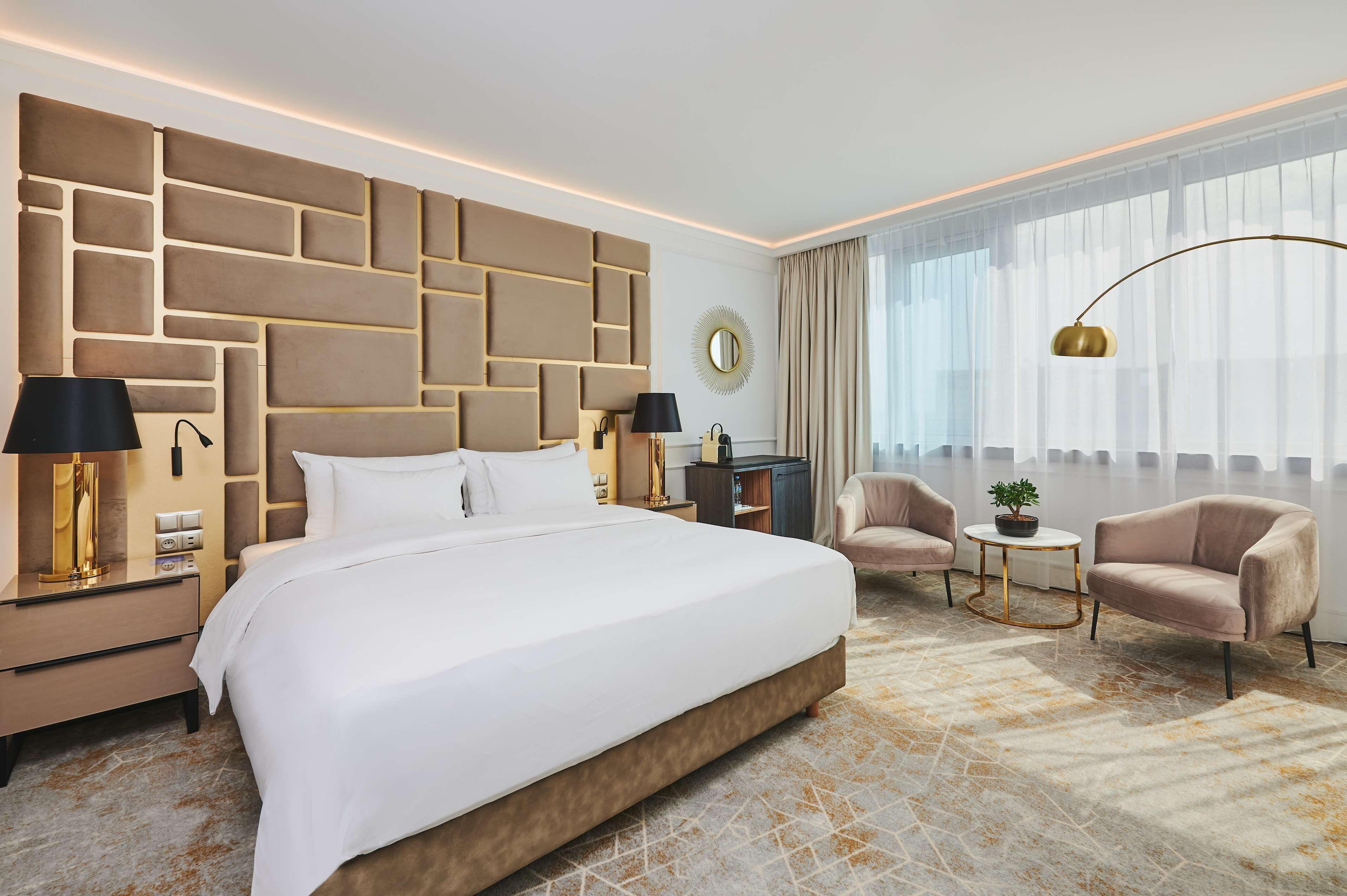 Una camera d'albergo con un letto grande, due comodini con lampade, uno specchio rotondo, un tavolino con una pianta e due poltrone.