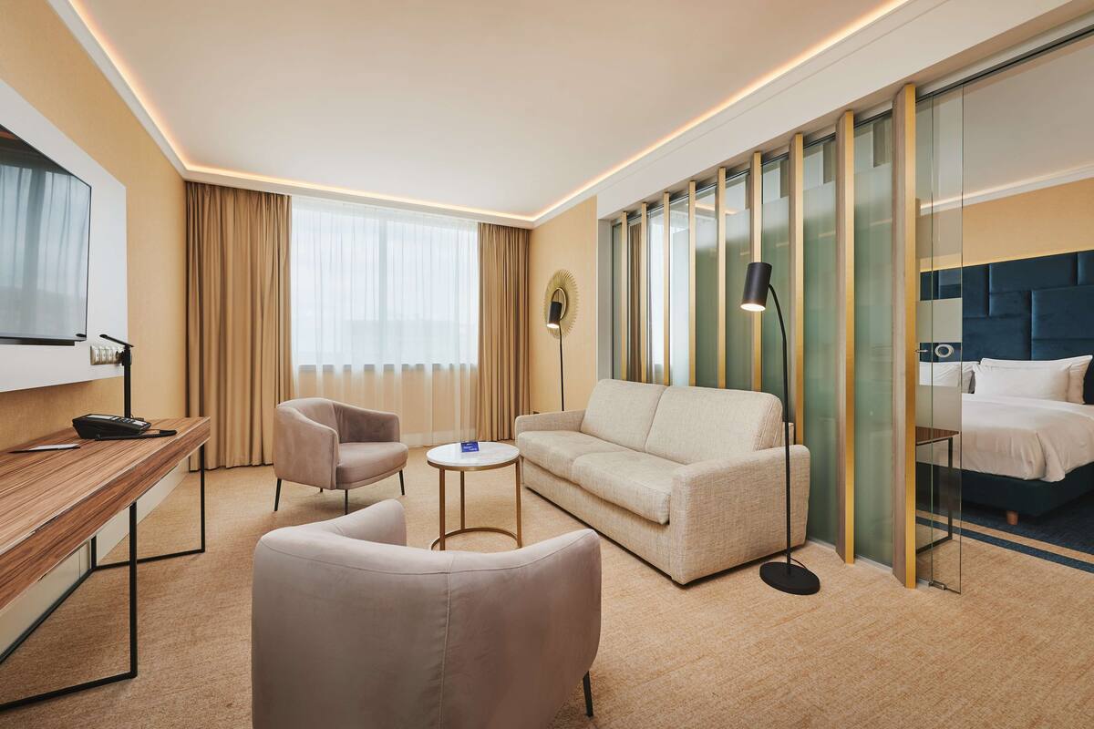 A hotel room with a sofa, two armchairs, a small round table, a desk with a phone, a TV, and a bed with a headboard.