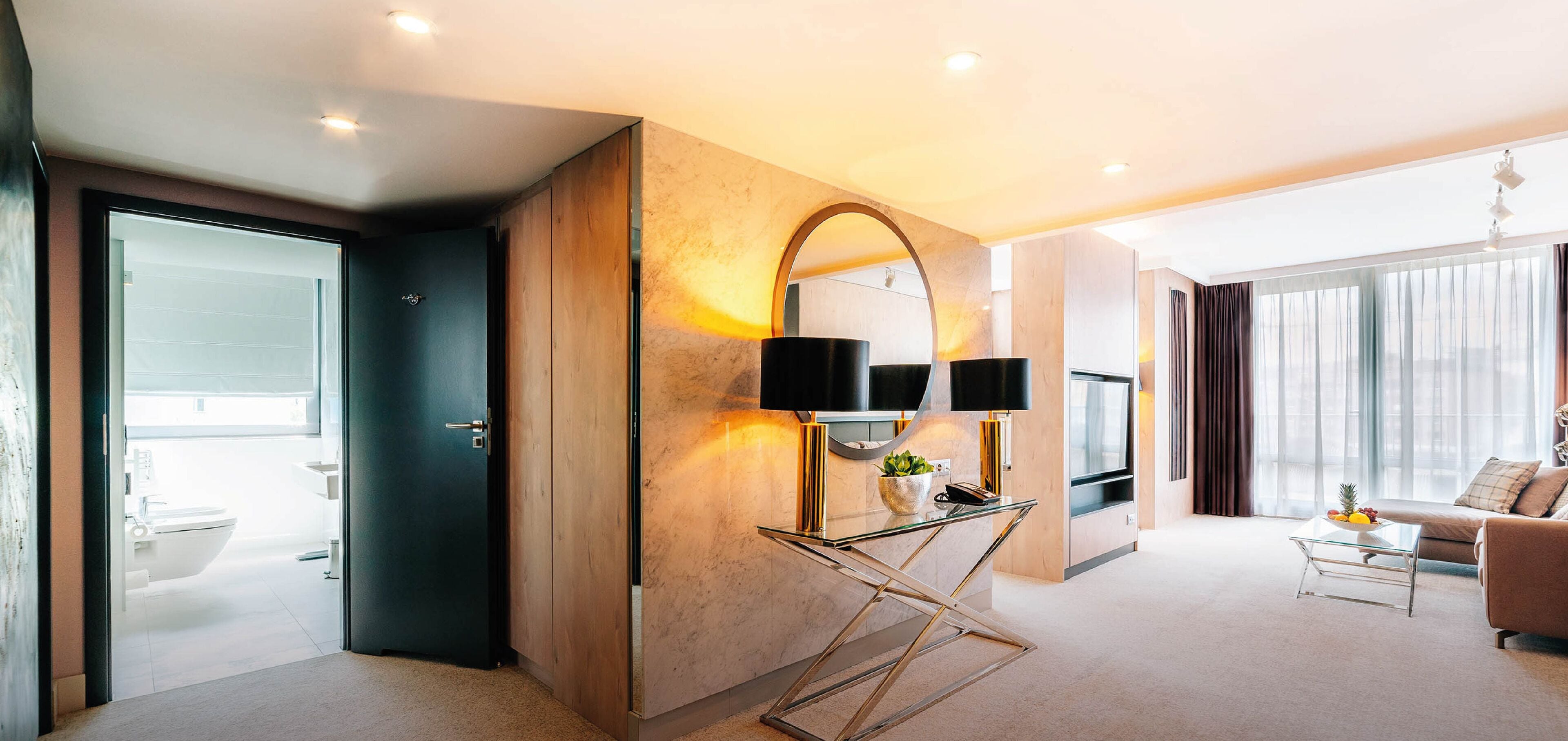 A modern hotel room with a glass partition separating the bathroom, a marble wall with a mirror and lamps, a sofa, and a coffee table with fruit.