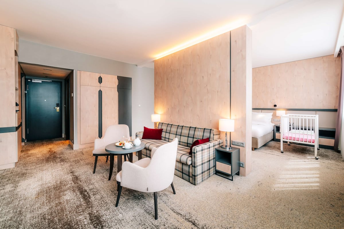 A hotel room with a sofa, armchair, and a crib.
