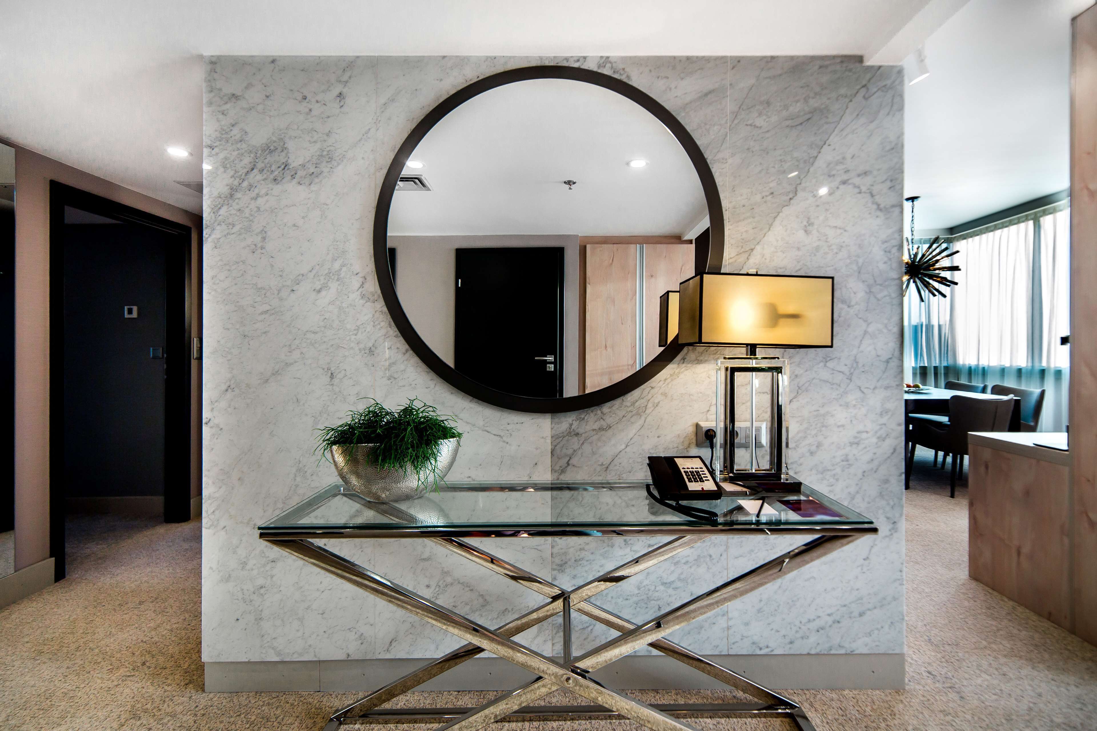 Um interior moderno com uma mesa de console de vidro, um espelho redondo, um vaso com planta, uma luminária e um telefone.
