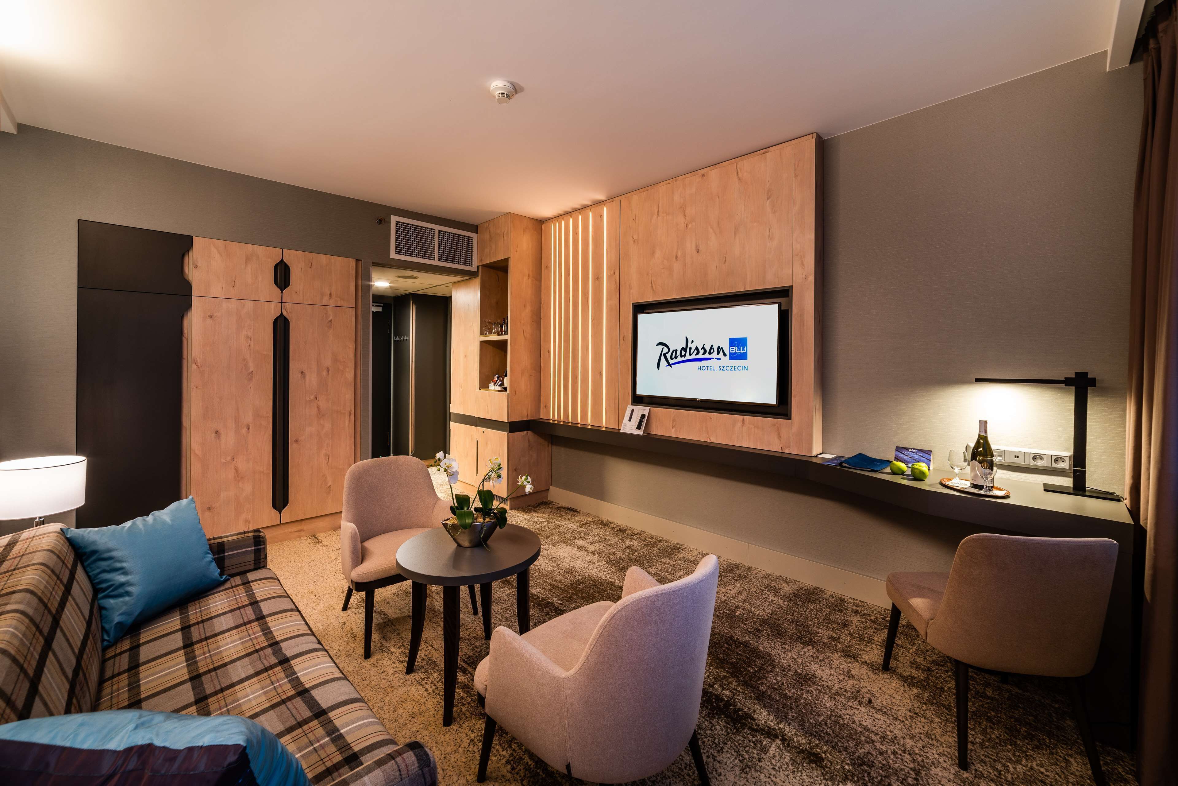 A modern hotel room with a living area featuring a sofa, two armchairs, a small round table, and a TV mounted on the wall.