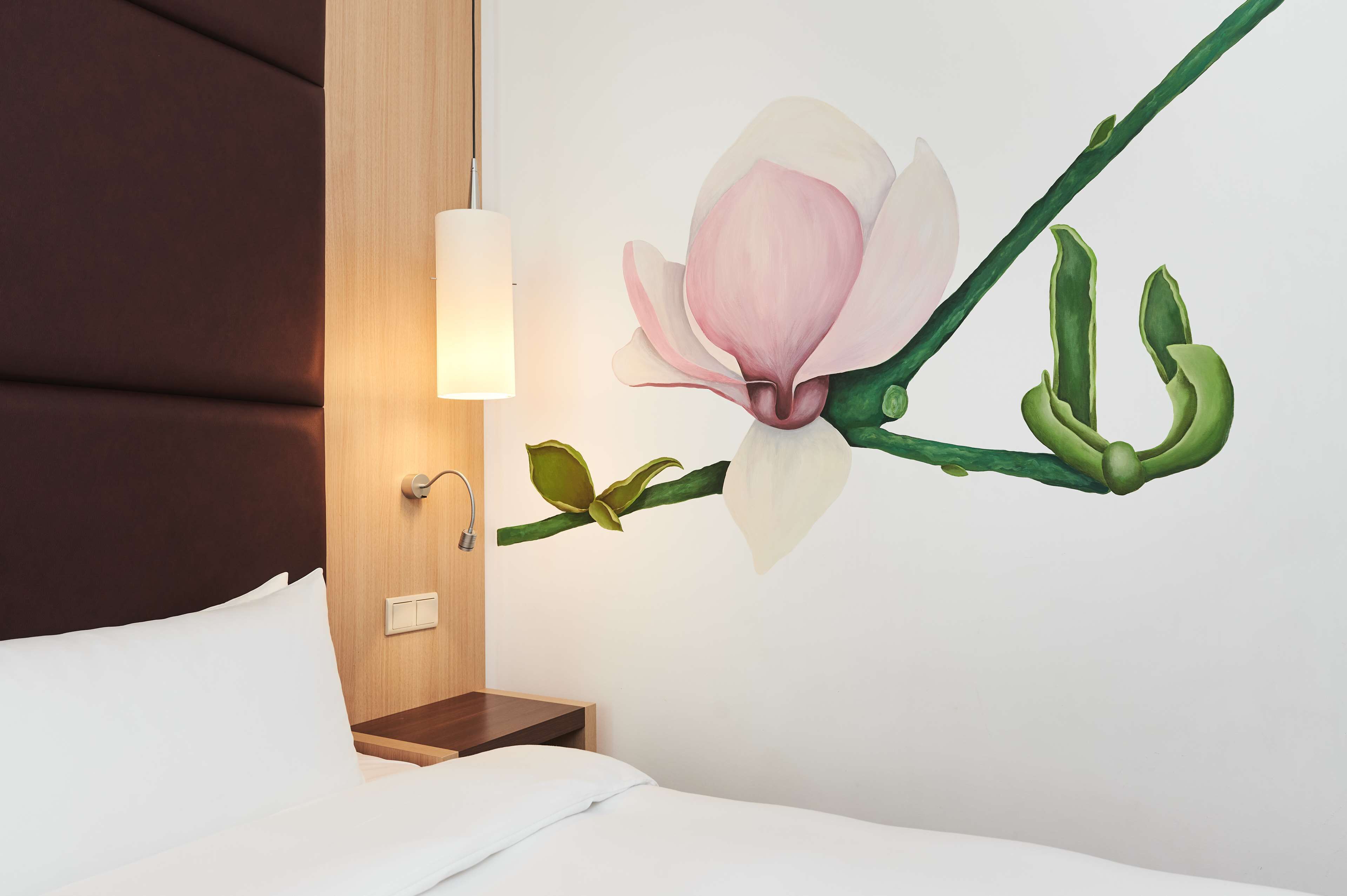 A hotel room with a bed, a nightstand, a wall-mounted lamp, and a floral mural.