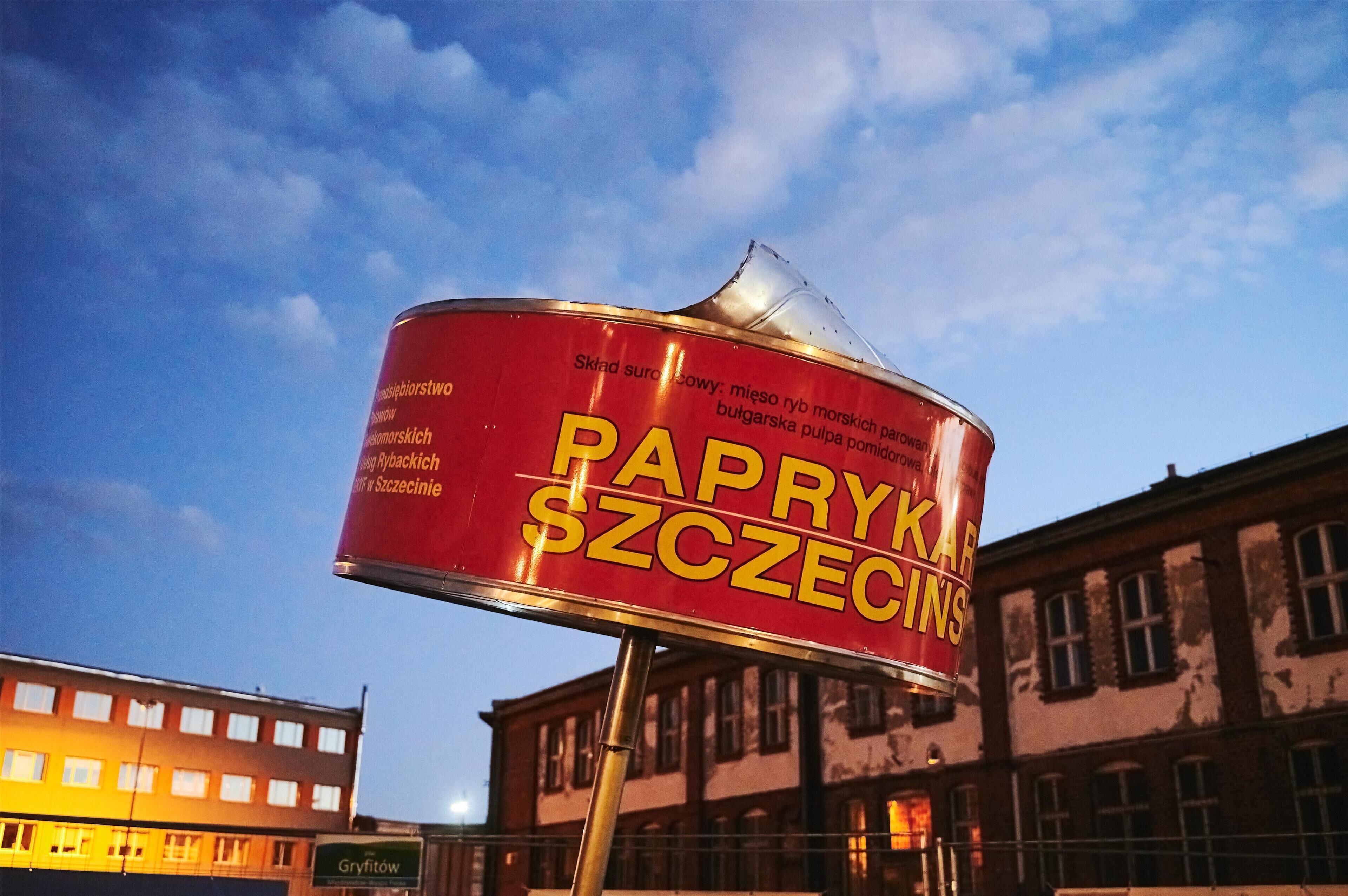 A red can with yellow text advertising 'PAPRYKA SZCZECINSKI' against a backdrop of buildings and a clear sky.