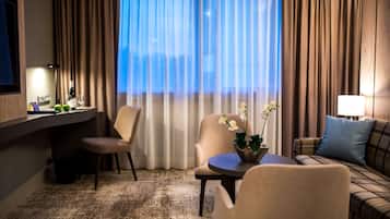 Una habitación de hotel con escritorio, dos sillas, un sofá, una mesita con flores, televisor y ventana con cortinas.