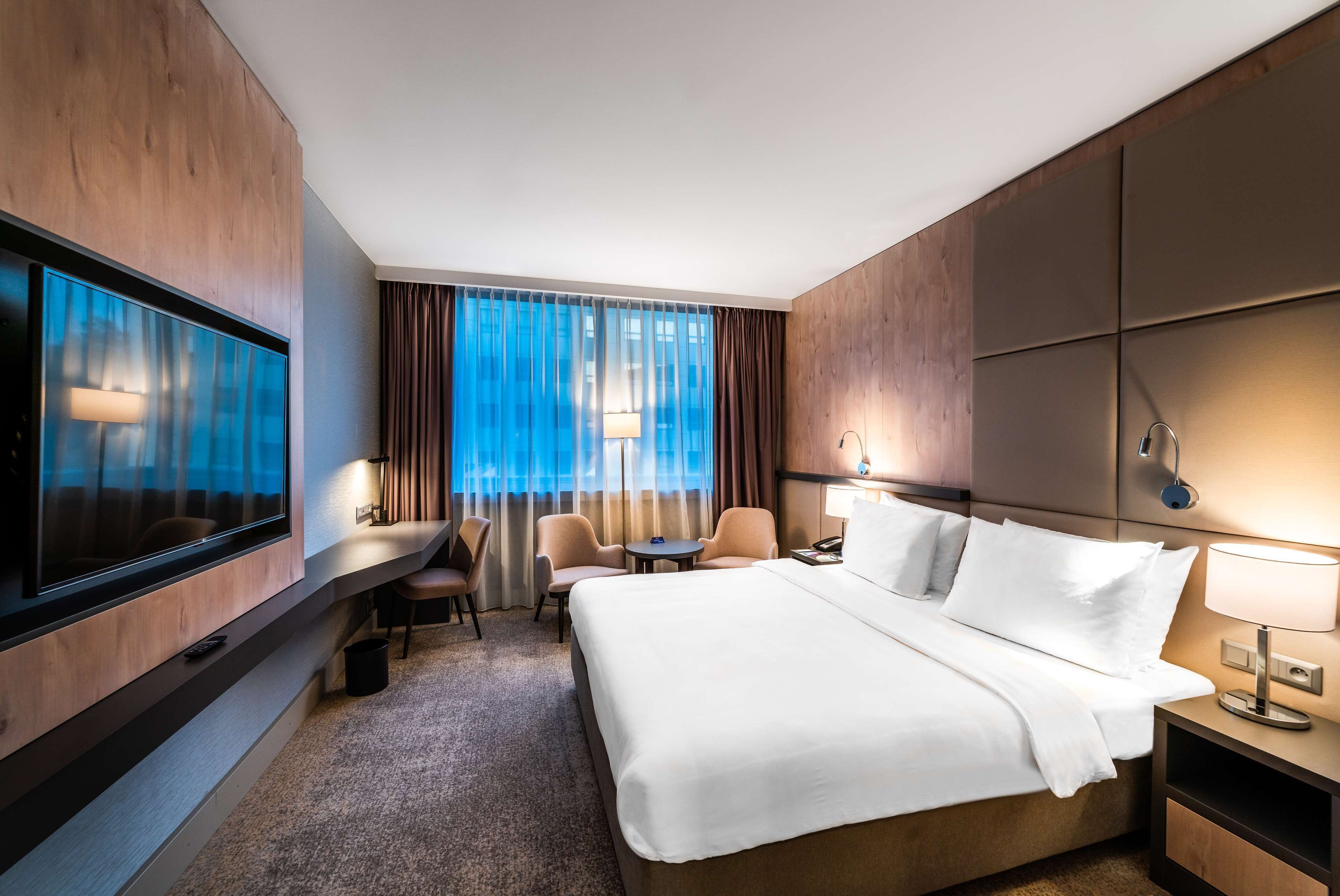 Ein Hotelzimmer mit einem großen Bett, einem Schreibtisch mit Stuhl, einem Fernseher und einem Fenster mit Vorhängen.