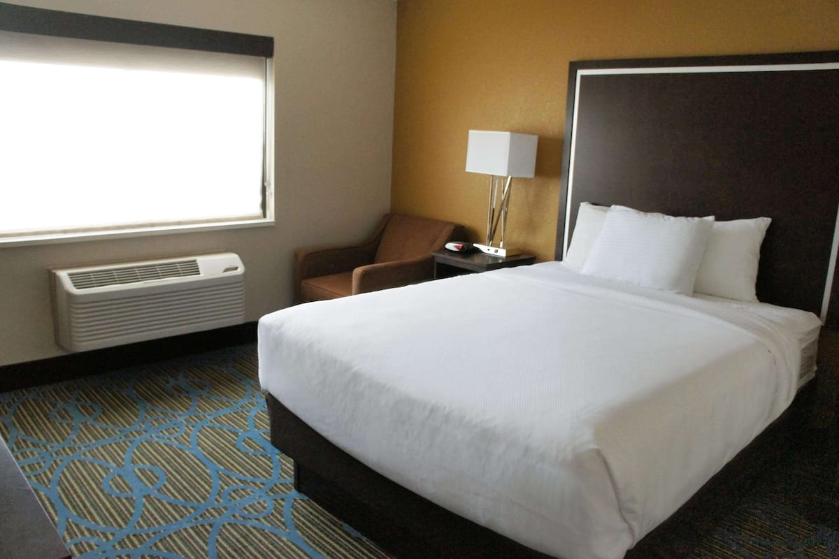 Ein Hotelzimmer mit einem großen Bett, einem Sessel, einem Schreibtisch mit Lampe und einem Fenster mit Jalousien.