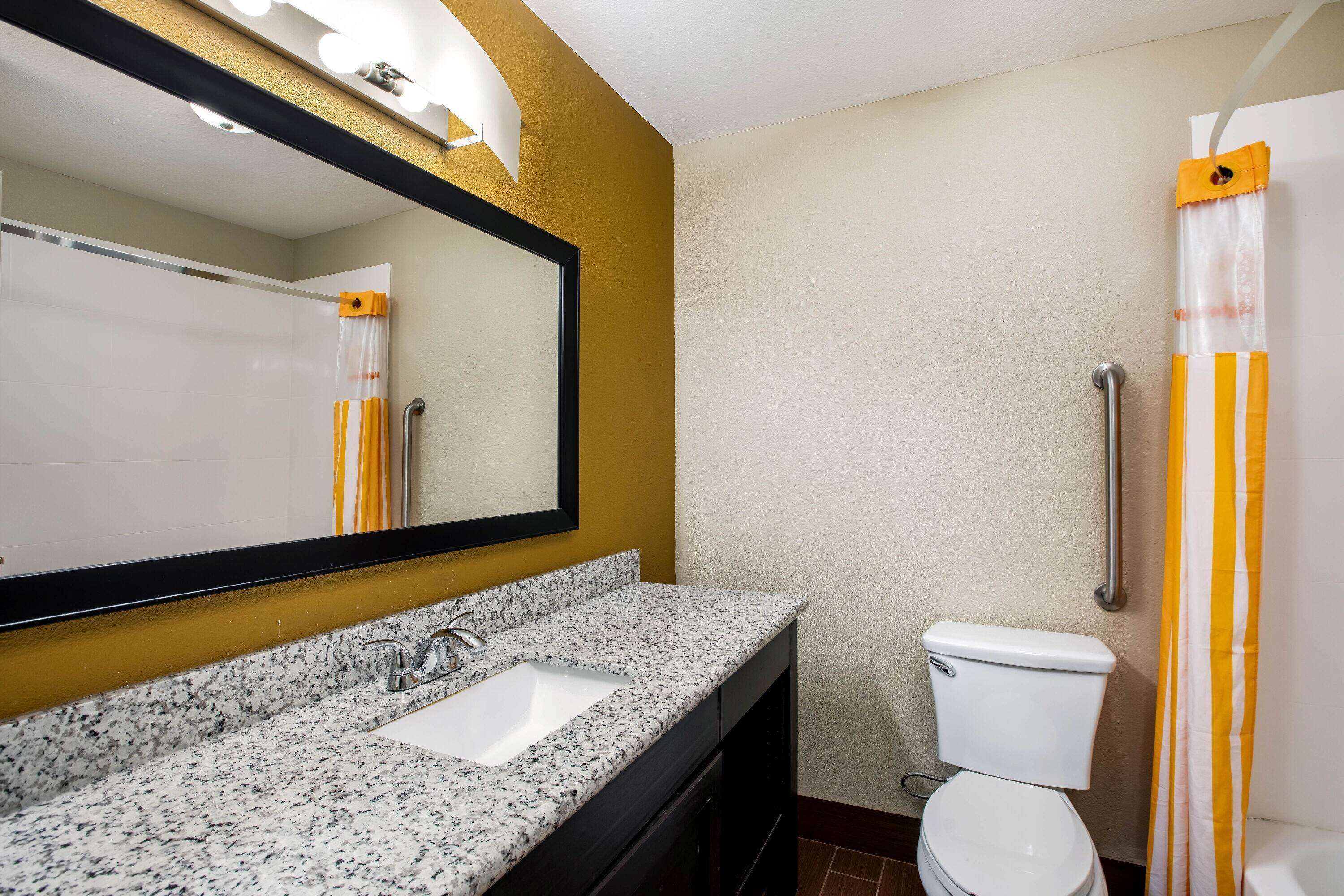 A bathroom with a granite countertop sink, a white toilet, and a mirror with lights above it.