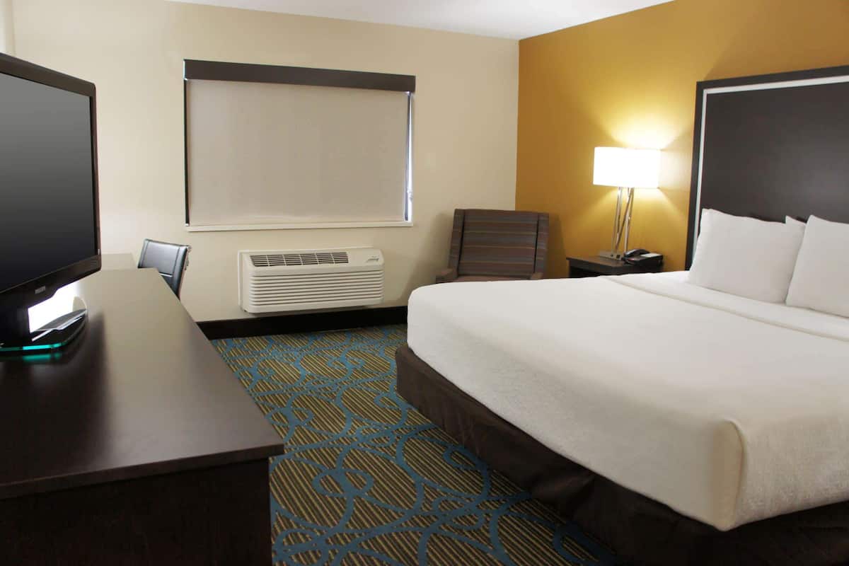 Ein Hotelzimmer mit Bett, Schreibtisch, Fernseher, Klimaanlage, Lampe und einem Fenster mit Jalousien.